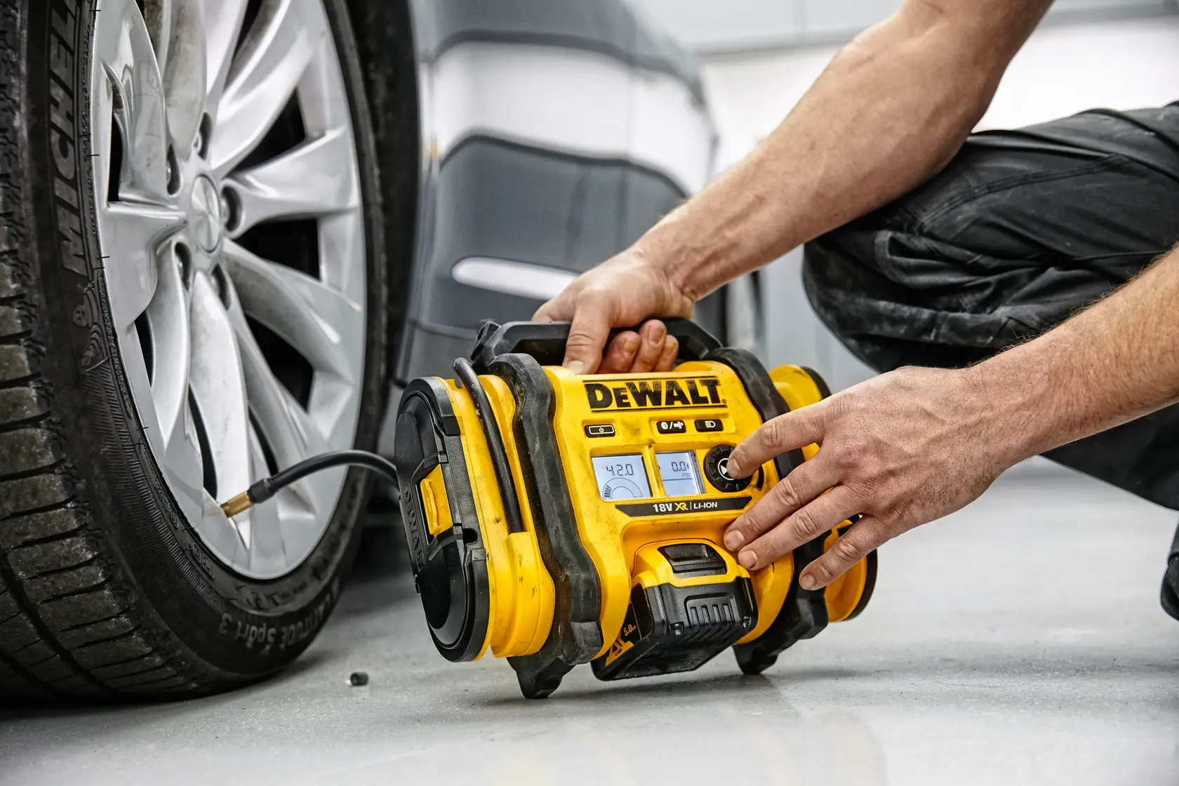 Eine Person verwendet einen gelben, tragbaren DEWALT-Kompressor, um den Reifen eines Autos aufzupumpen. Das Gerät ist mit dem Reifenventil verbunden und steht neben dem Fahrzeug auf dem Boden.