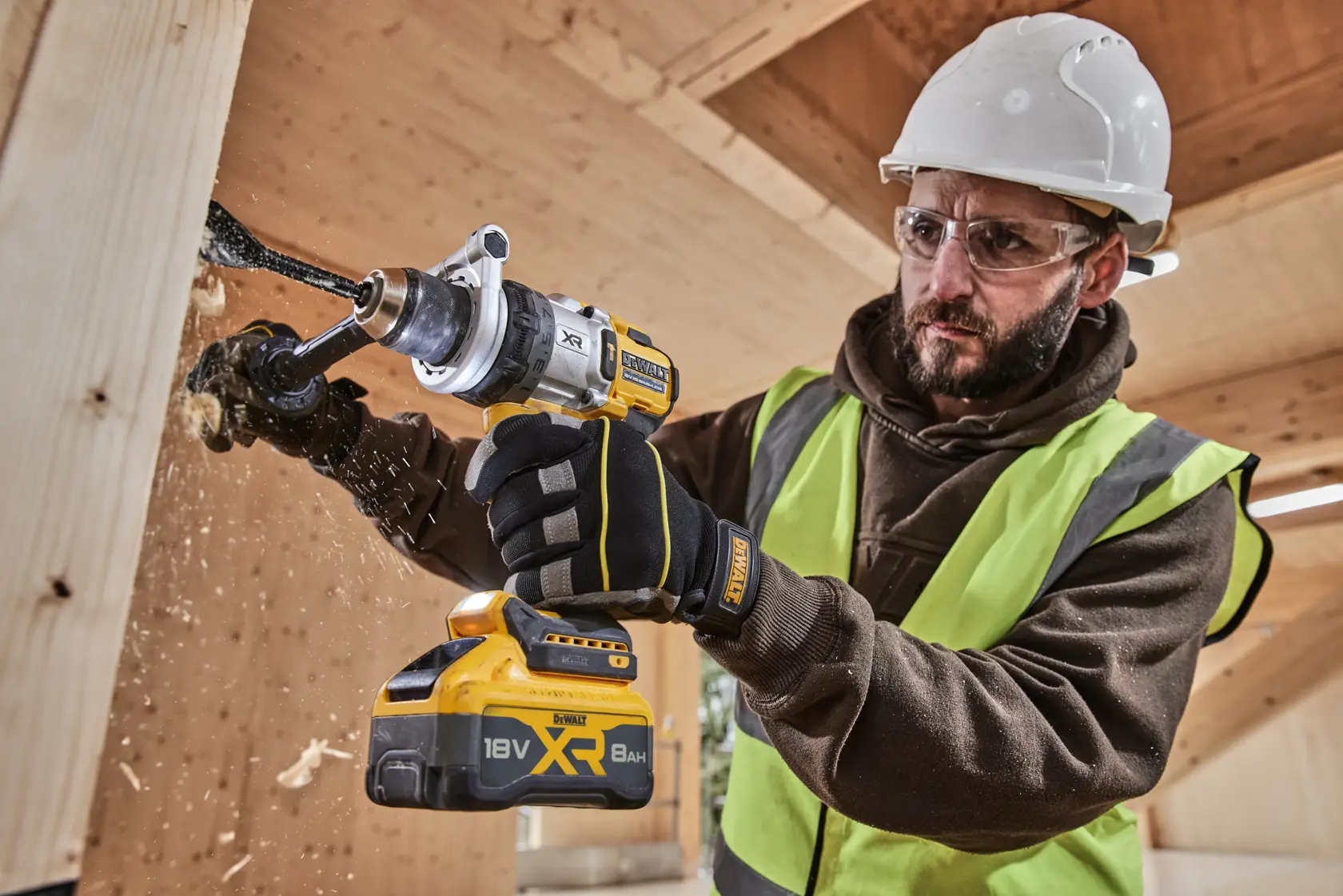 Trabajador perfora madera con DEWALT 18V XR, usando guantes, casco y chaleco en obra.
