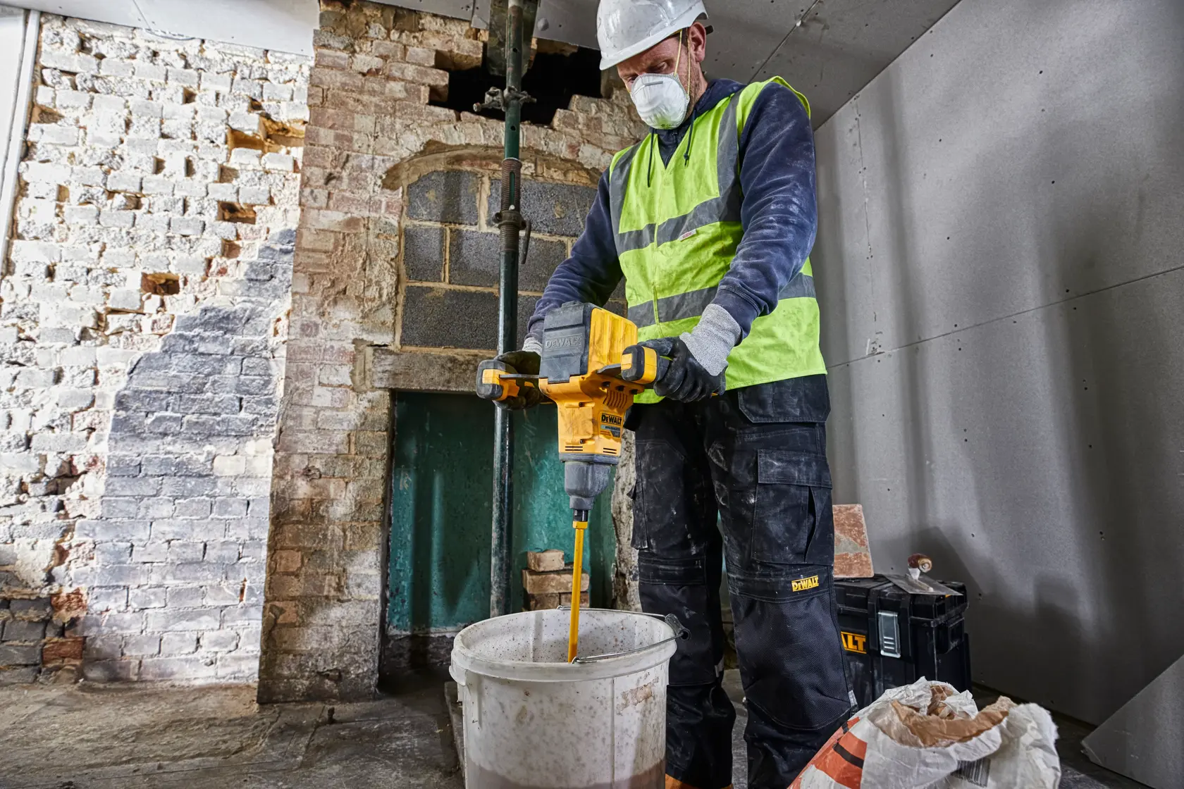Ein Bauarbeiter mit Warnweste und Schutzhelm bedient einen gelben DEWALT DCD240N Rührwerk, um Materialien in einem weißen Eimer in einem teilweise renovierten Raum zu mischen.