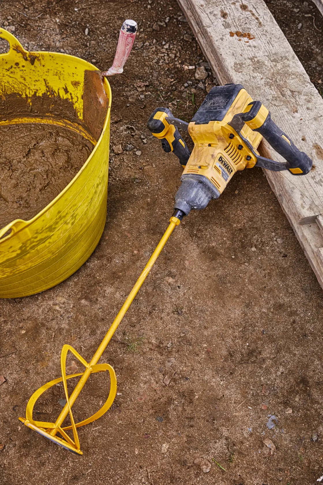 A DEWALT cordless mixer with a long yellow mixing paddle is lying on a construction site floor next to a yellow bucket containing mixed material and a trowel.