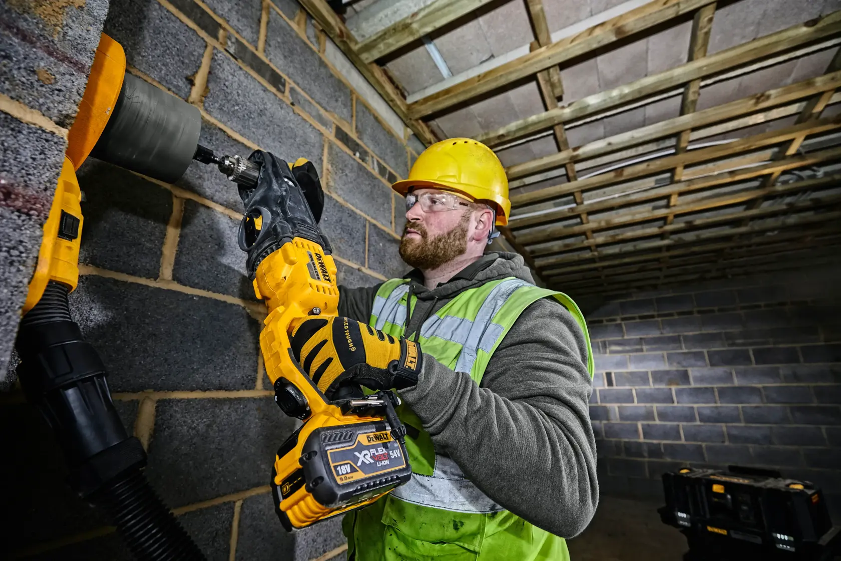 Eine Person mit gelbem Schutzhelm, Sicherheitshandschuhen und Warnweste verwendet einen kabellosen DEWALT-Bohrer (DCD470N), um ein Loch in eine Betonblockwand auf einer Baustelle zu bohren. Das Gesicht der Person ist verschwommen.