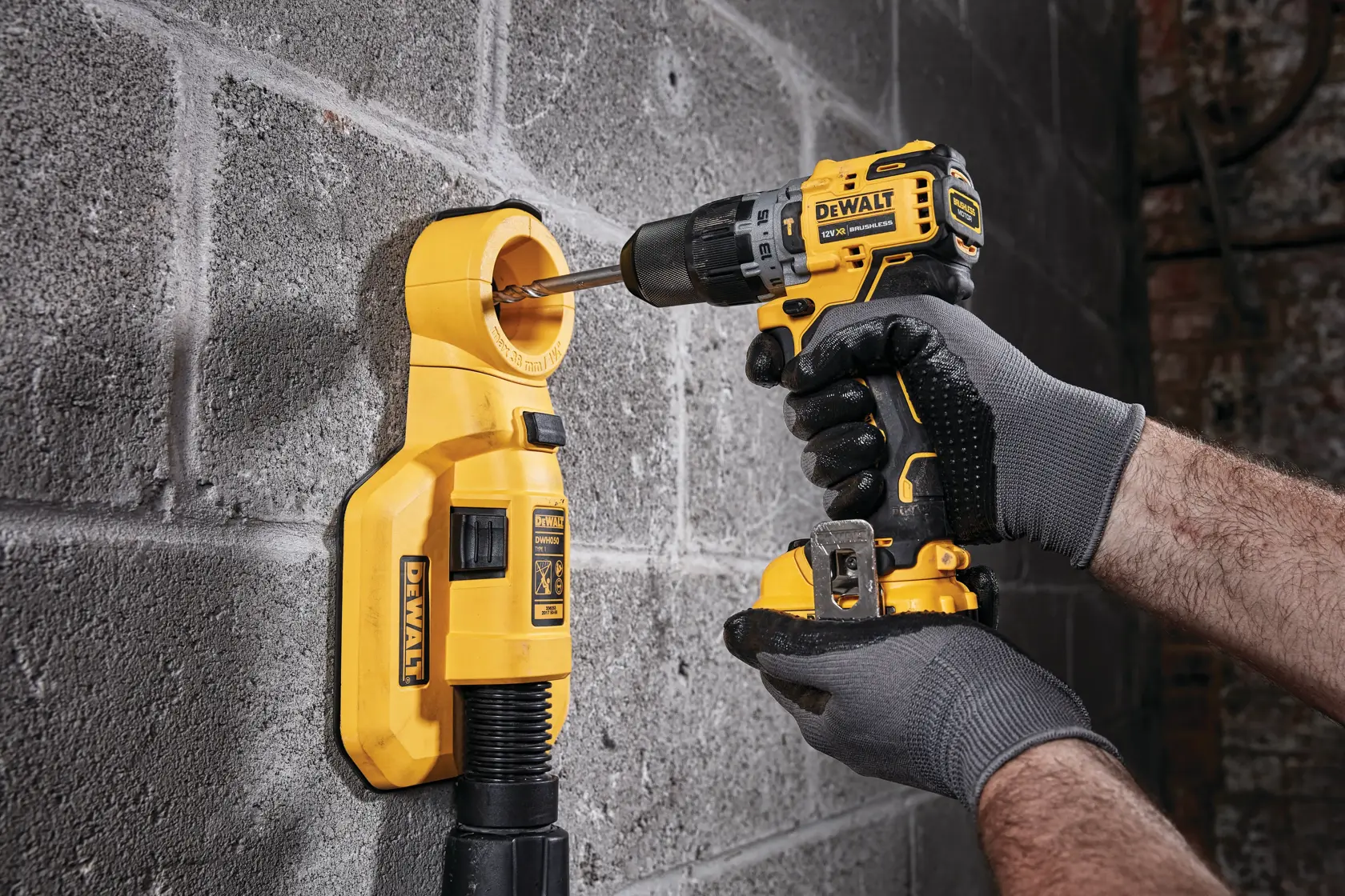 Una persona con guanti grigi utilizza un trapano a batteria giallo DEWALT per forare una parete in cemento, con un aspiratore di polvere giallo collegato per raccogliere i detriti.
