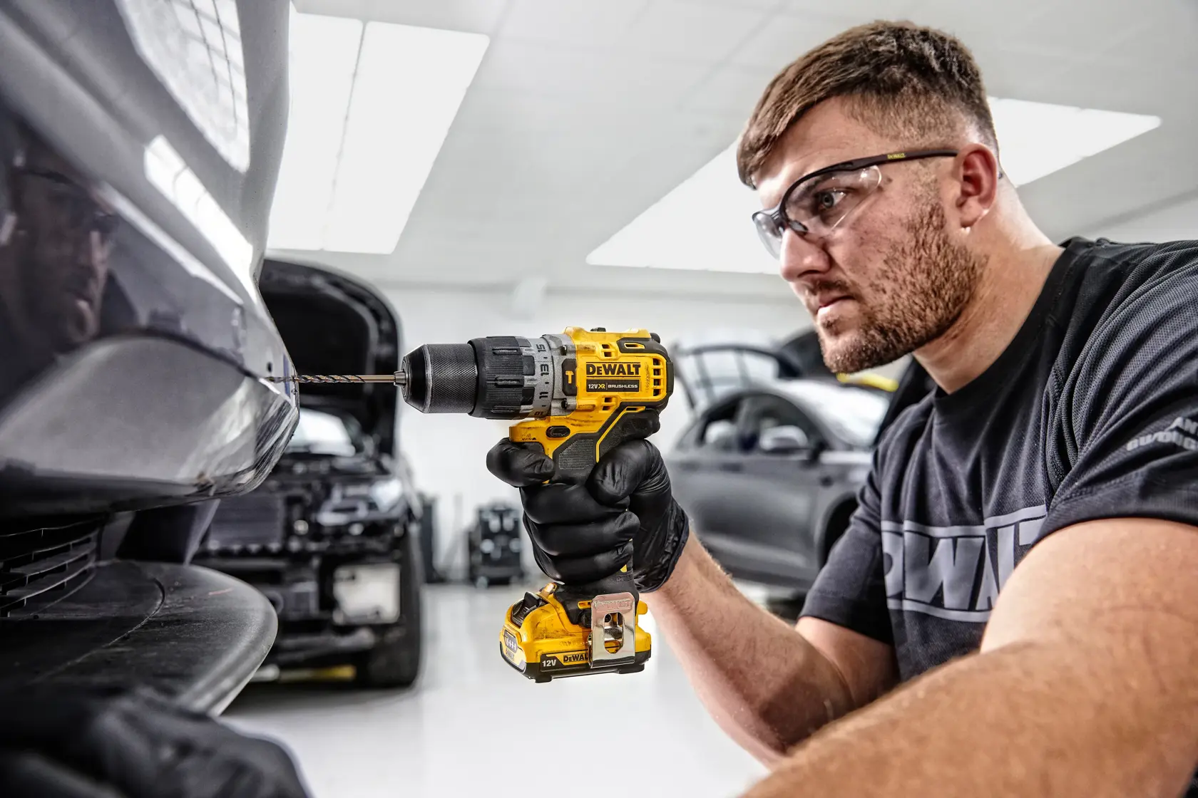 Una persona con guanti neri utilizza un trapano a batteria giallo DEWALT DCD706D2 per forare il paraurti di un’auto in un’officina automobilistica. Il volto della persona è sfocato e sullo sfondo sono visibili diverse auto.