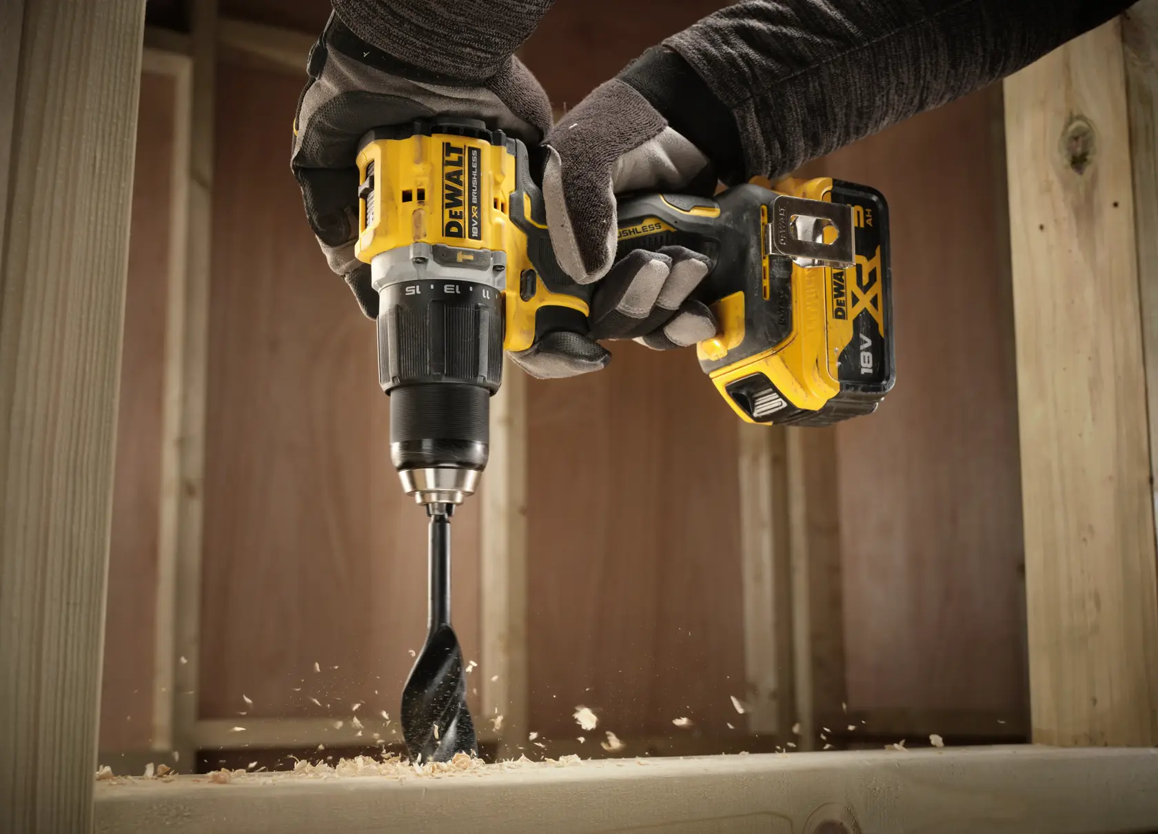 Taladro inalámbrico DEWALT perfora madera, virutas vuelan, sujetado por manos enguantadas en obra.