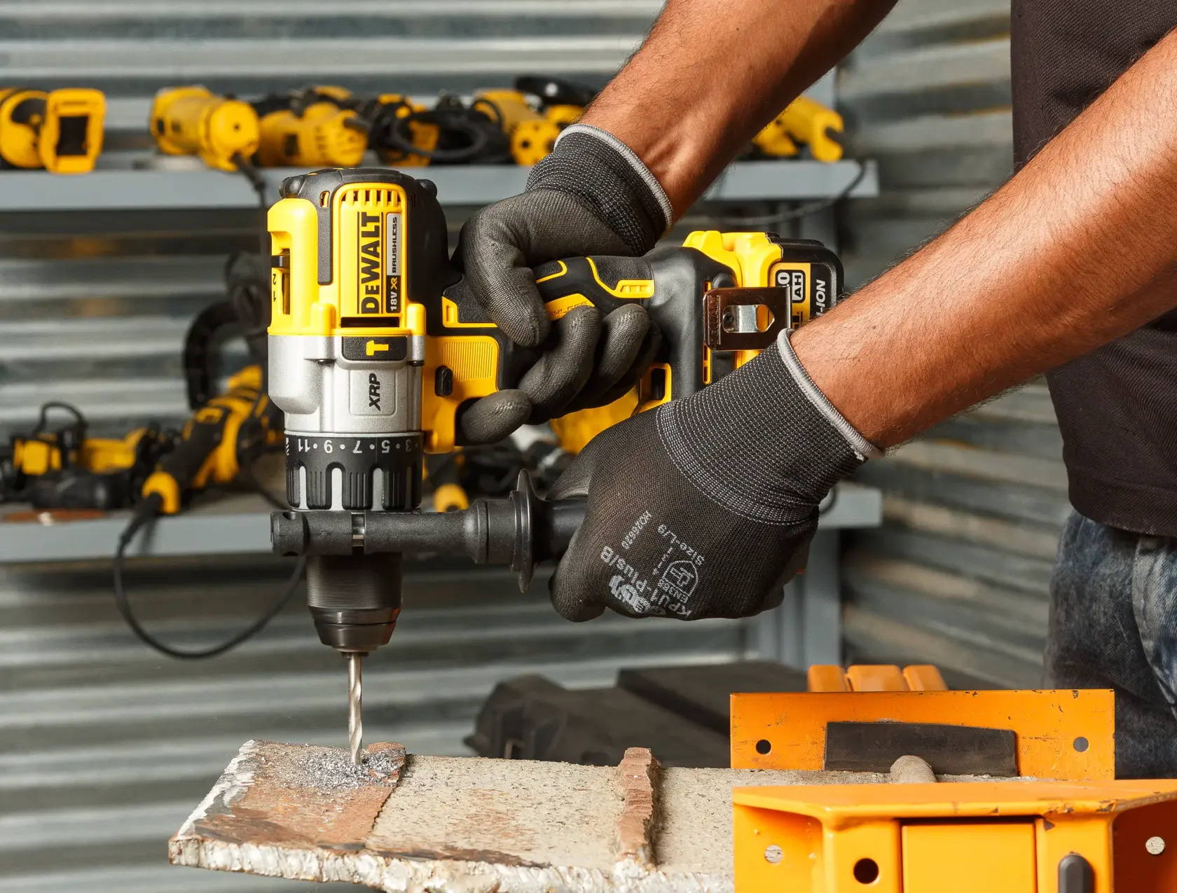 Persona perfora metal con taladro inalámbrico DEWALT y guantes. Hay herramientas en estantes cercanos.