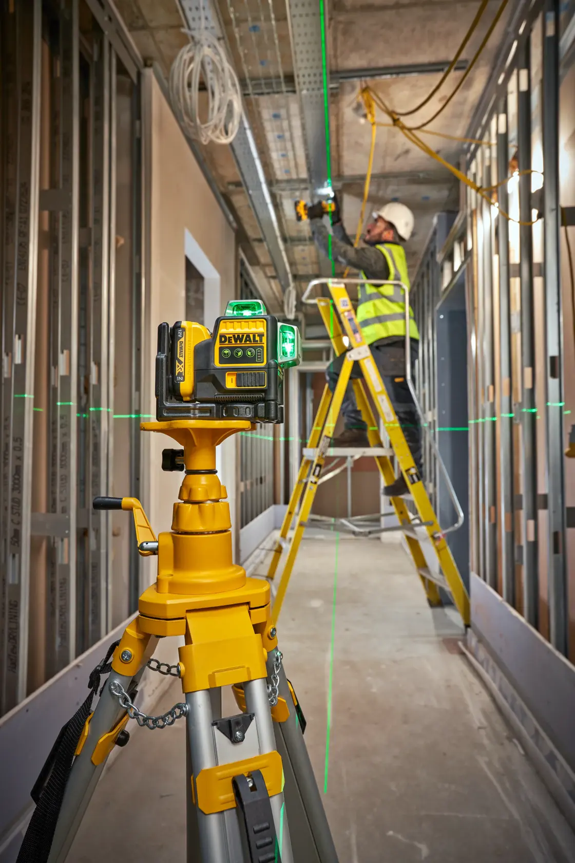 Nivel láser DEWALT sobre trípode proyecta líneas verdes; trabajador usa escalera en zona de construcción.