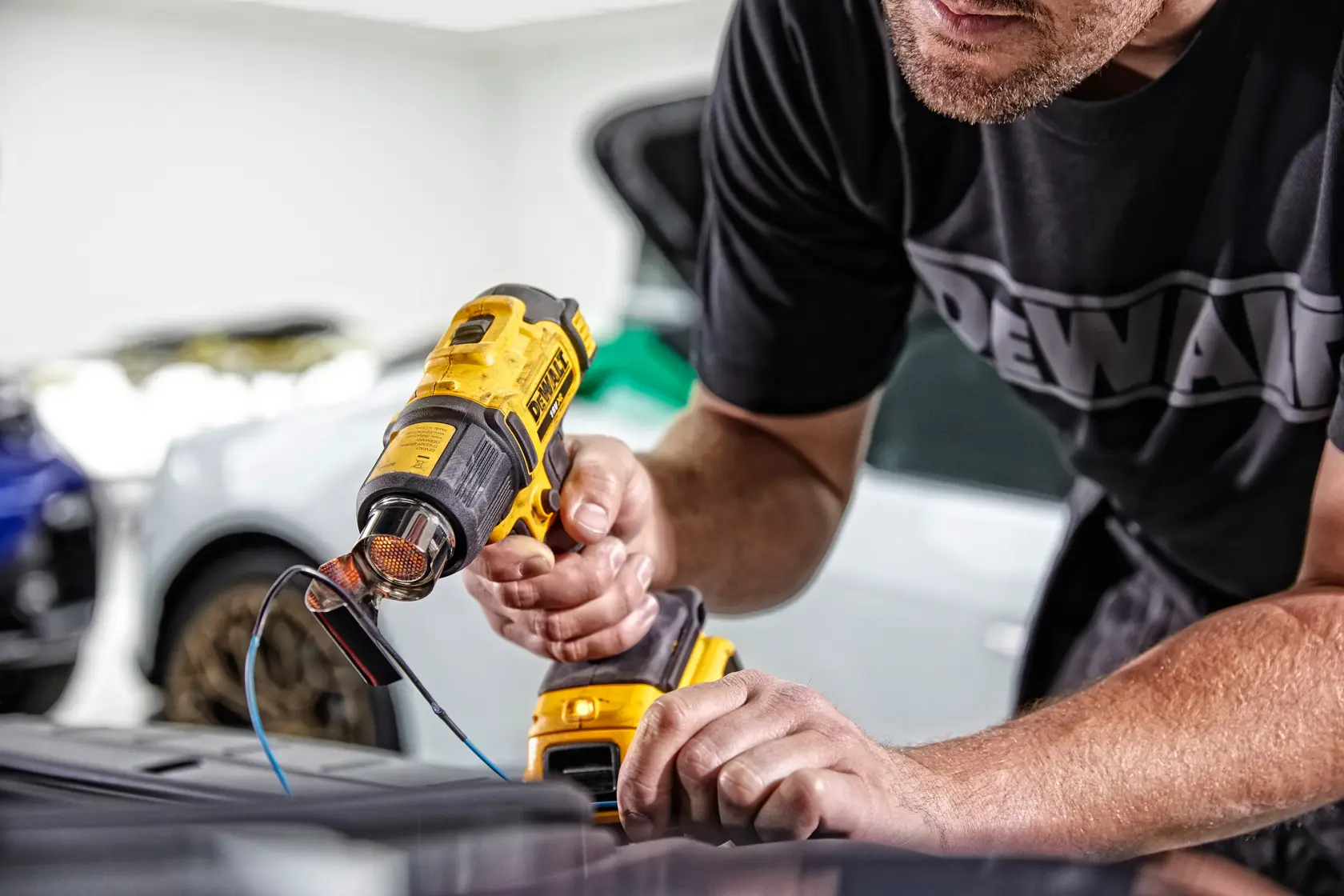 Una persona utiliza una pistola de calor inalámbrica DEWALT DCE530N para trabajar sobre una superficie, con cables y autos visibles en el fondo.