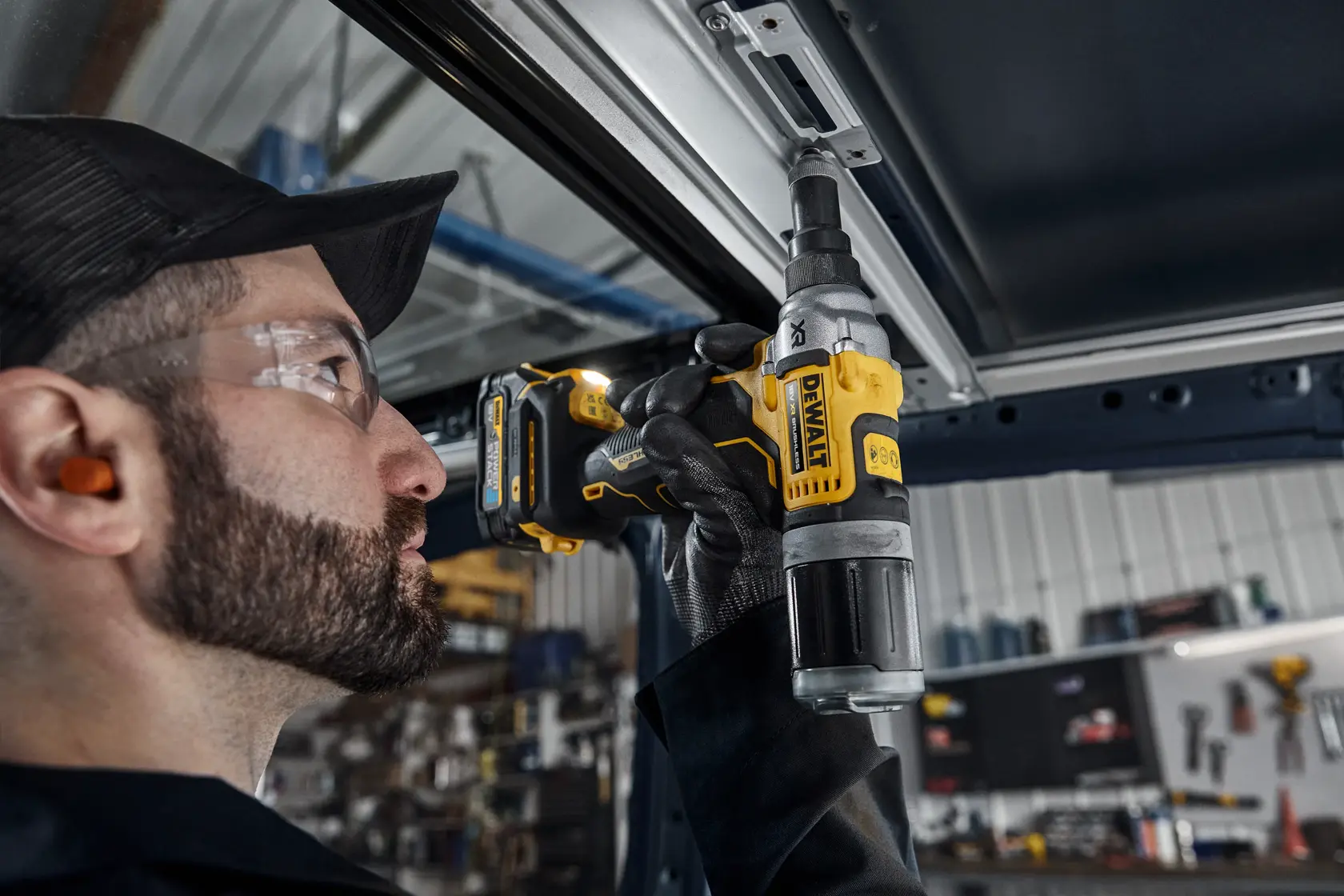 Una persona con protección auditiva y guantes usa una herramienta inalámbrica DEWALT DCF414 para fijar o taladrar un marco de metal, con un taller visible al fondo.