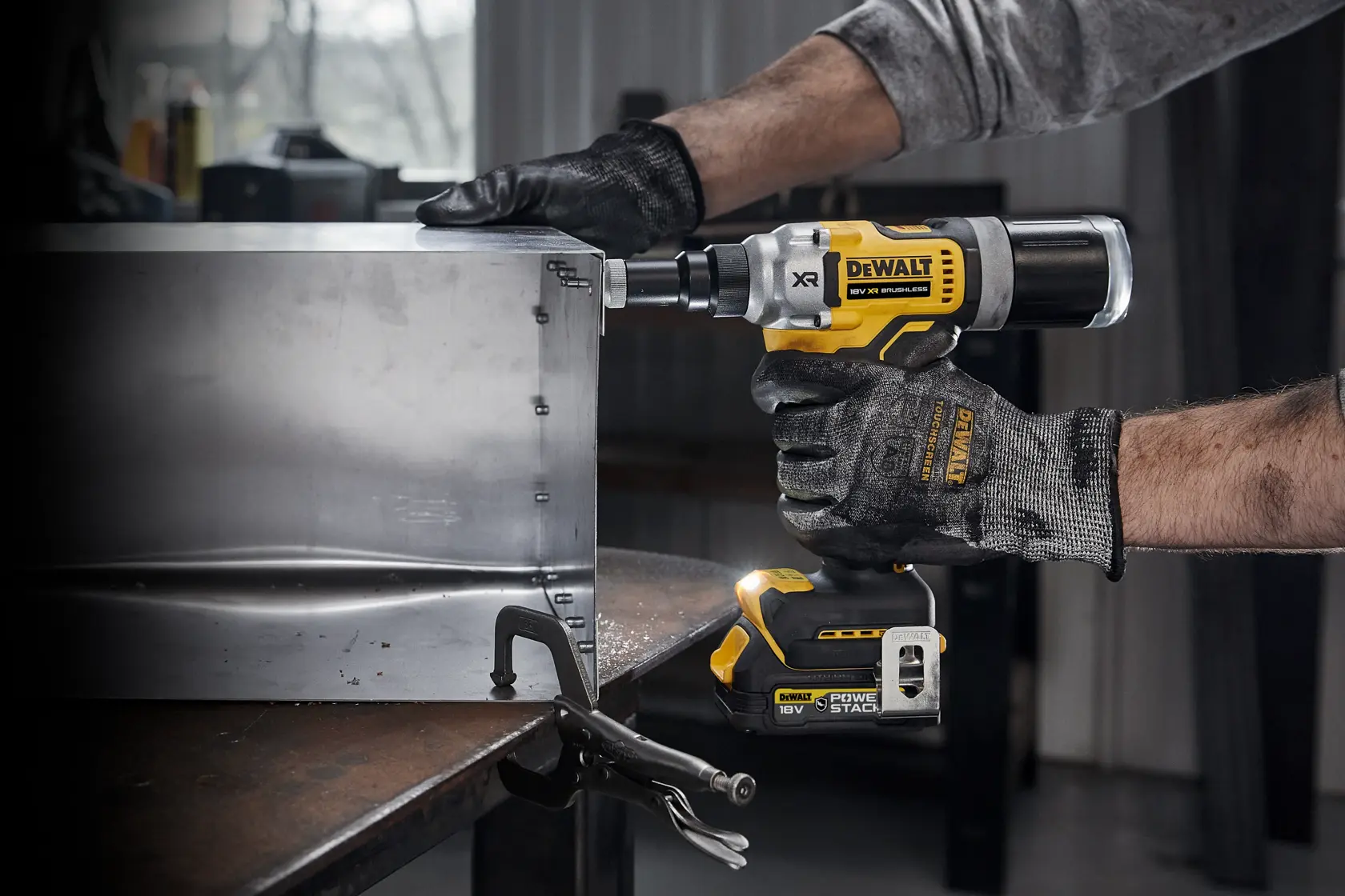 Una persona con guantes utiliza una remachadora inalámbrica DEWALT 18V XR (modelo DCF414) para sujetar láminas de metal en una mesa de trabajo en un taller.