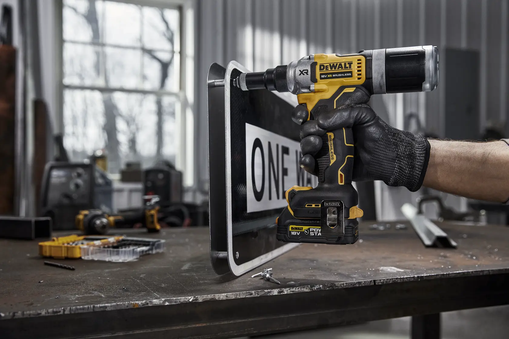 Una persona con guante negro usa una herramienta eléctrica DEWALT DCF414E2GT amarilla y negra para perforar o fijar una señal de tráfico en un banco de trabajo metálico en un taller industrial.