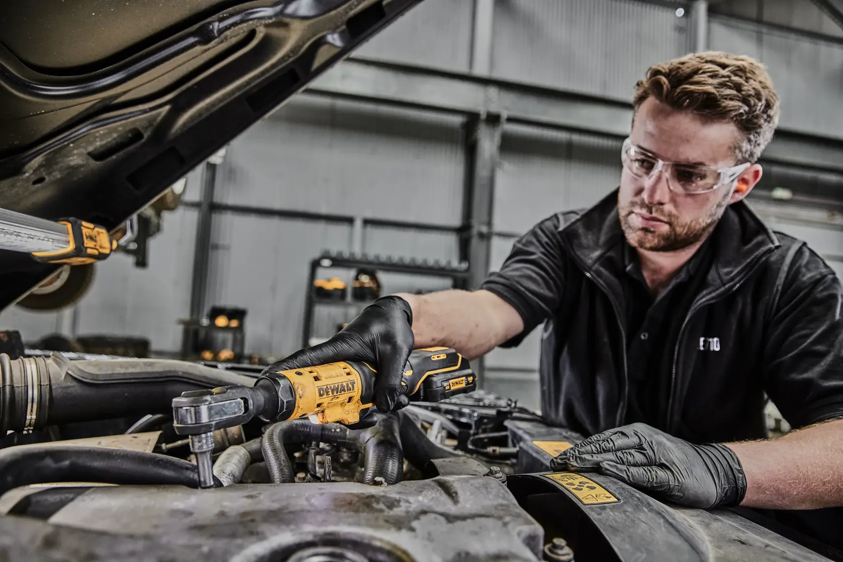Eine Person mit schwarzen Handschuhen benutzt ein kabelloses Knarrenwerkzeug von DEWALT, um an einem Automotor in einer Werkstatt zu arbeiten. Das Gesicht der Person ist verpixelt.