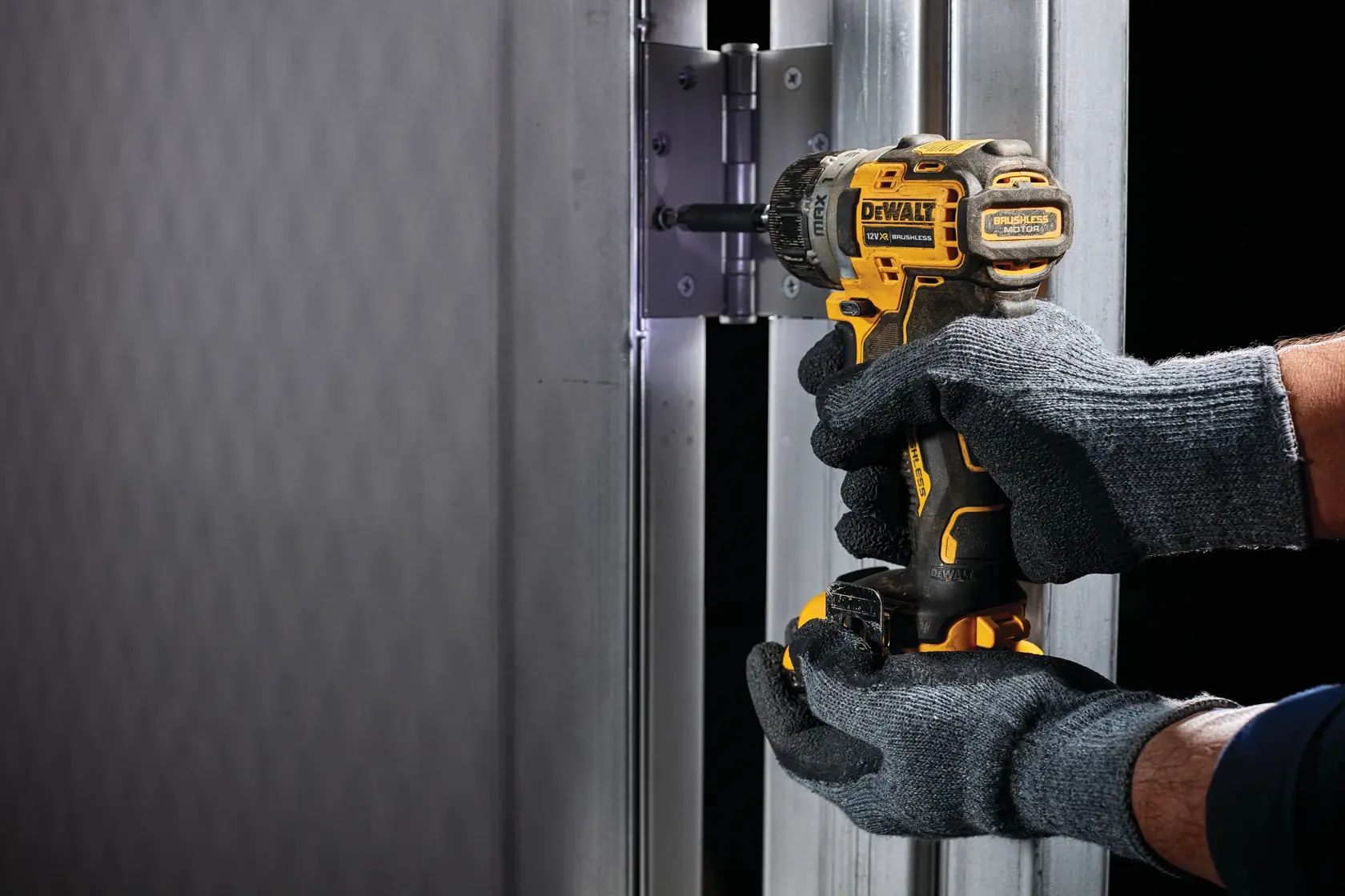 Manos con guantes usan un destornillador DEWALT DCF601N inalámbrico para apretar tornillos de una bisagra metálica.