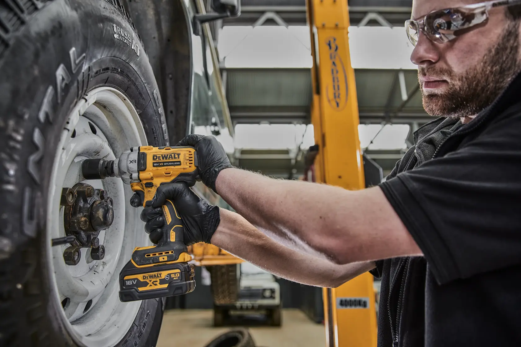 Una persona con guanti neri utilizza un avvitatore a impulsi senza fili DEWALT DCF891P2G per stringere i bulloni di una ruota di veicolo in un'officina.