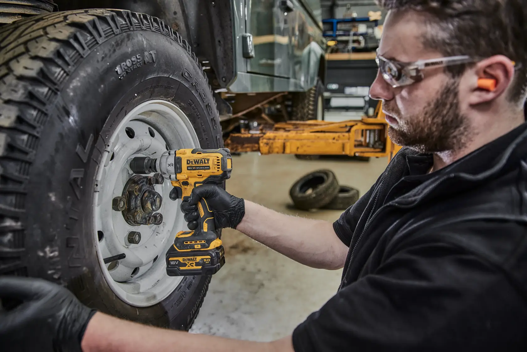 Una persona con i guanti utilizza un avvitatore a impulsi senza fili DEWALT (modello DCF891P2G) per lavorare sui dadi di una ruota di veicolo in un’officina. Il viso della persona è sfocato per motivi di privacy.