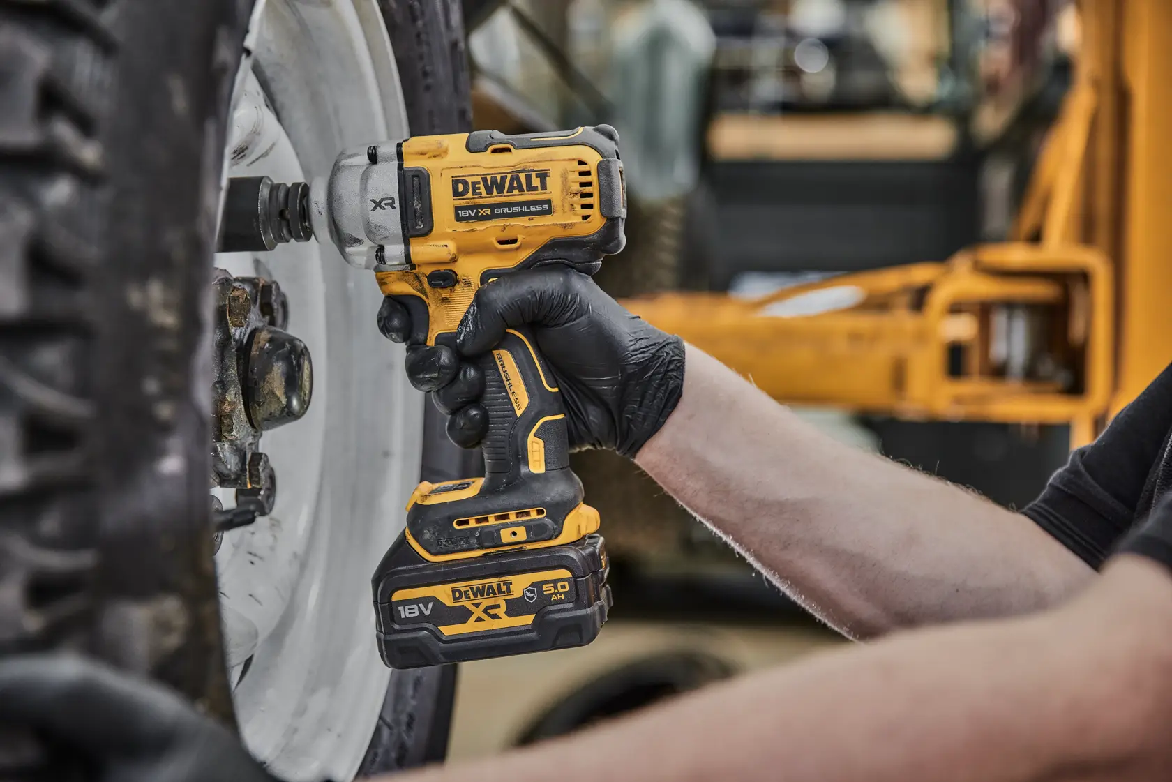 Una persona con guanti neri utilizza una chiave a impulsi senza fili gialla DEWALT (modello DCF891P2G, 18V XR) per lavorare sui bulloni di una grande ruota di veicolo.