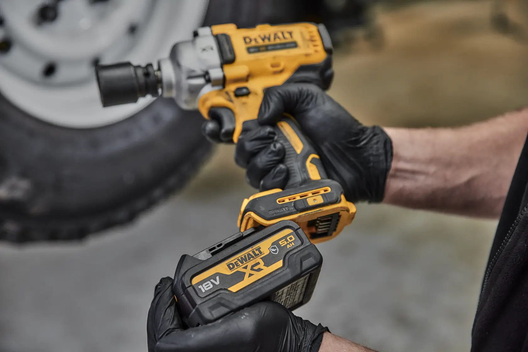 Una persona con guanti neri tiene in mano un avvitatore a impulsi cordless giallo e nero DEWALT e una batteria al litio DEWALT XR 18V 5,0 Ah. Sullo sfondo si vede un pneumatico sfocato.