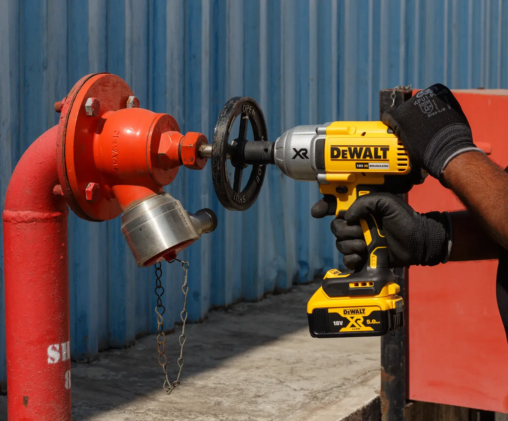Person uses a DEWALT cordless impact wrench to turn a valve on a red outdoor hydrant beside a blue wall.