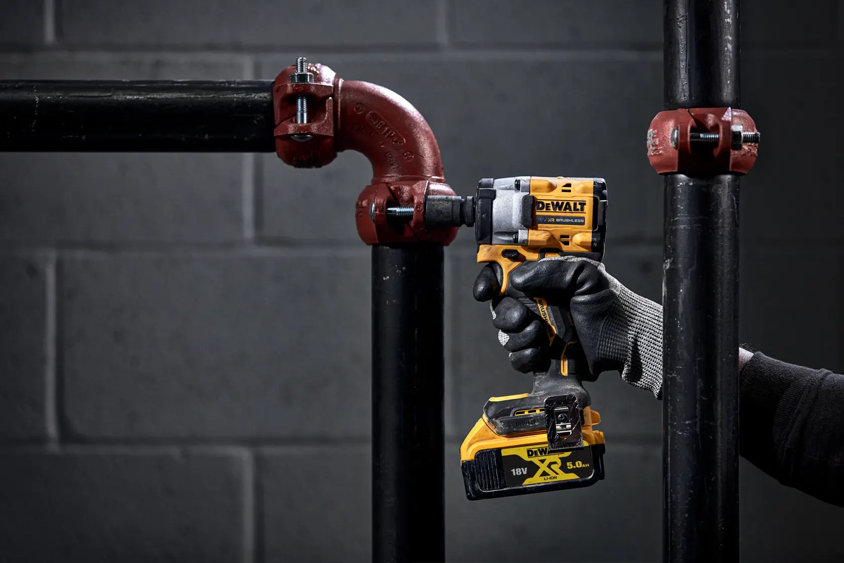 A person wearing black gloves is using a yellow DEWALT cordless power tool to tighten a metal pipe fitting.