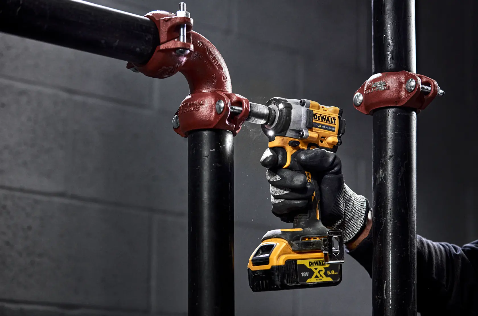 A person wearing gloves uses a DEWALT DCF921NT cordless impact wrench to tighten or loosen a metal pipe fitting in an industrial setting.