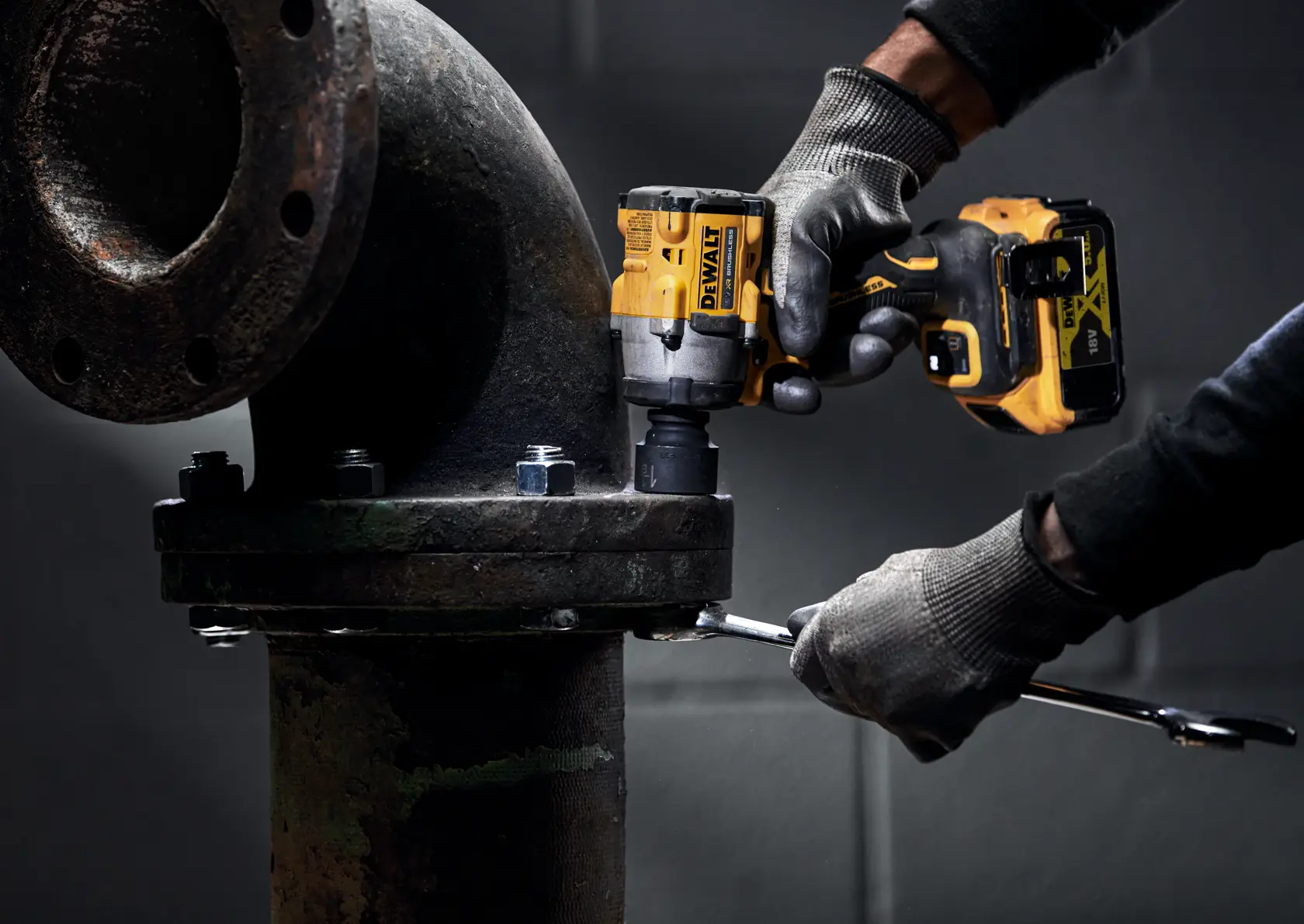 A person wearing gloves uses a DEWALT cordless impact wrench to tighten bolts on a large industrial pipe.