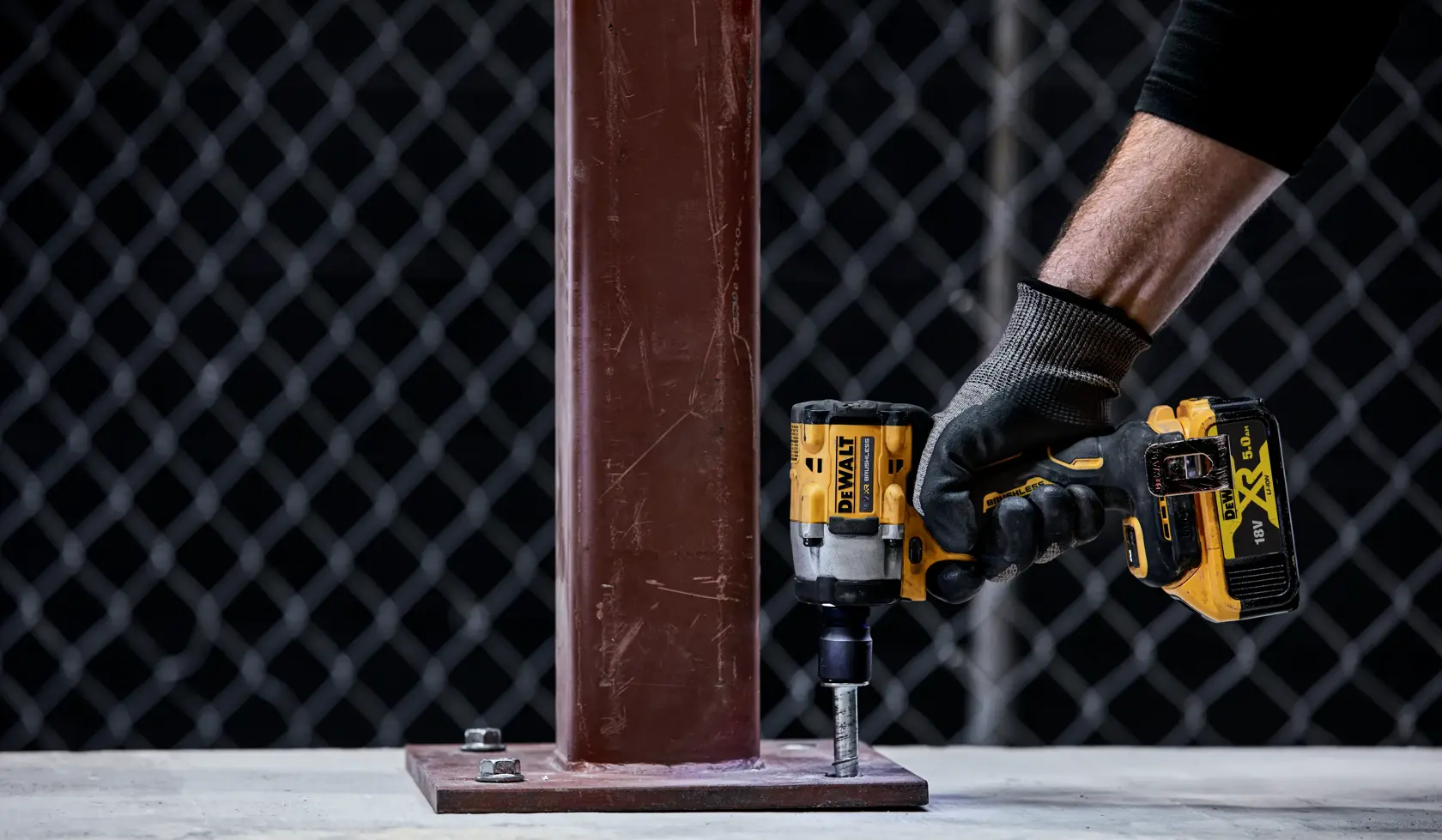 A person wearing a work glove uses a DEWALT DCF921NT cordless impact wrench to tighten a bolt at the base of a metal column.