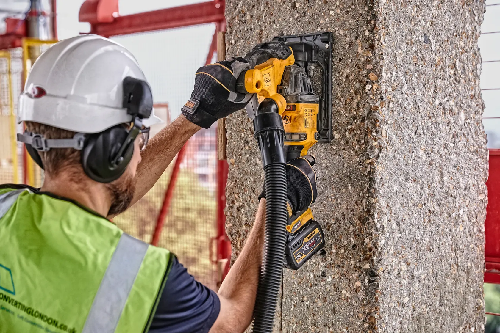 Un trabajador con casco de seguridad, guantes y chaleco reflectante utiliza una herramienta inalámbrica DEWALT para cortar o lijar un pilar de hormigón. La herramienta está equipada con una manguera de extracción de polvo para mayor seguridad y limpieza.