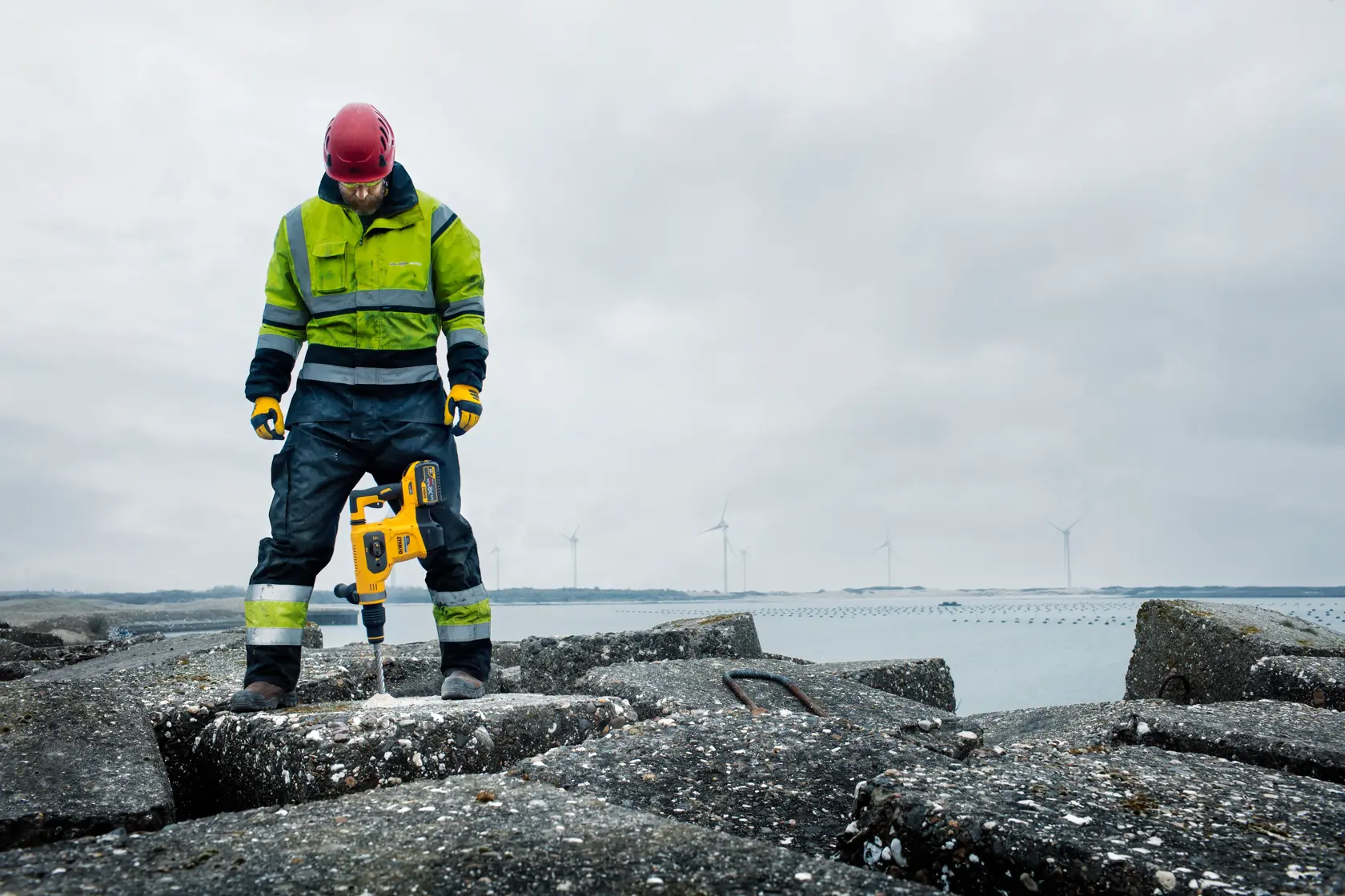 Una persona con ropa de trabajo de alta visibilidad y casco rojo utiliza un martillo rotativo DEWALT amarillo (SKU: DCH481N) para perforar concreto en un sitio de construcción al aire libre junto al agua, con turbinas eólicas visibles al fondo.