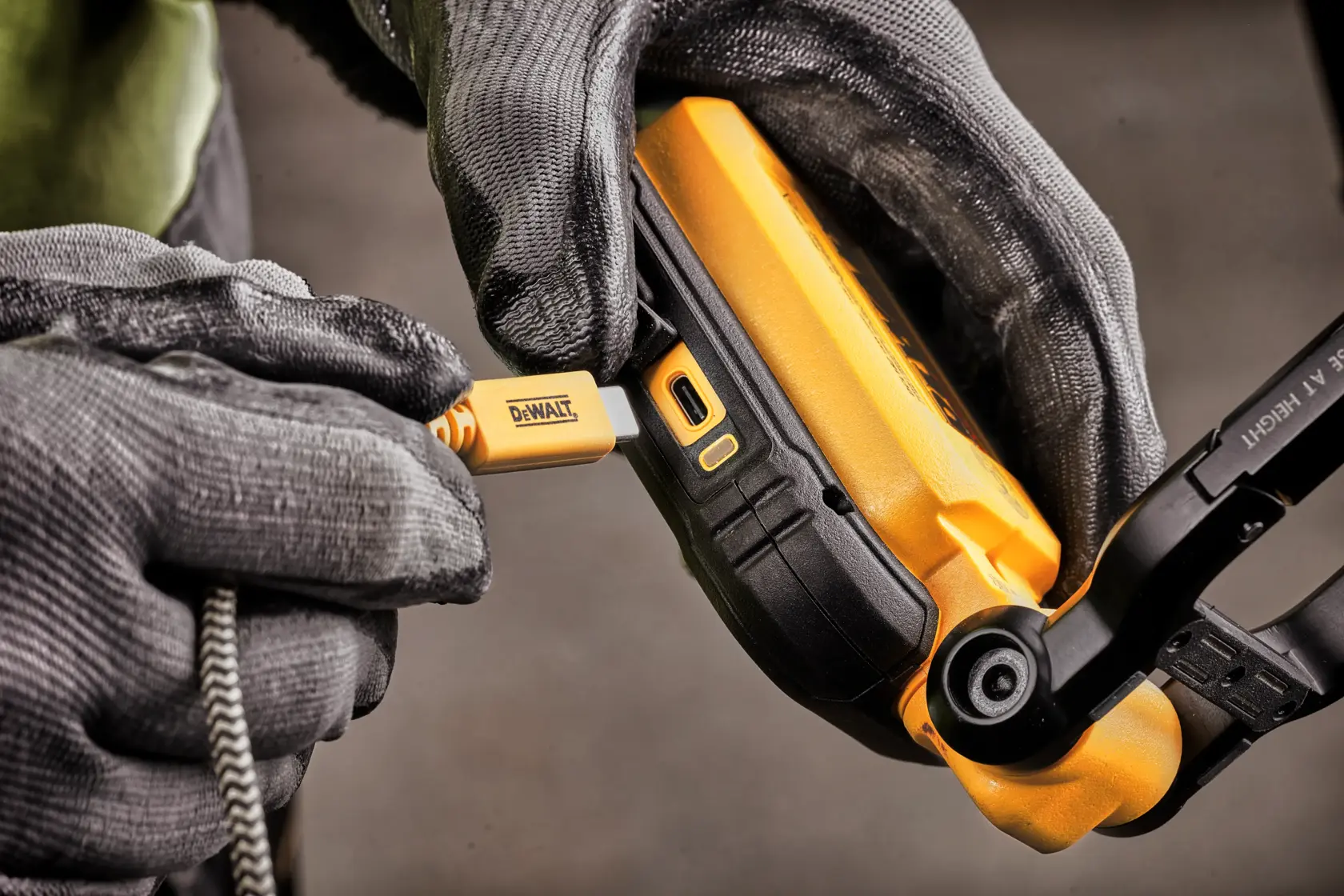 Close-up image of a person wearing gloves plugging a DEWALT charging cable into a DEWALT portable device.