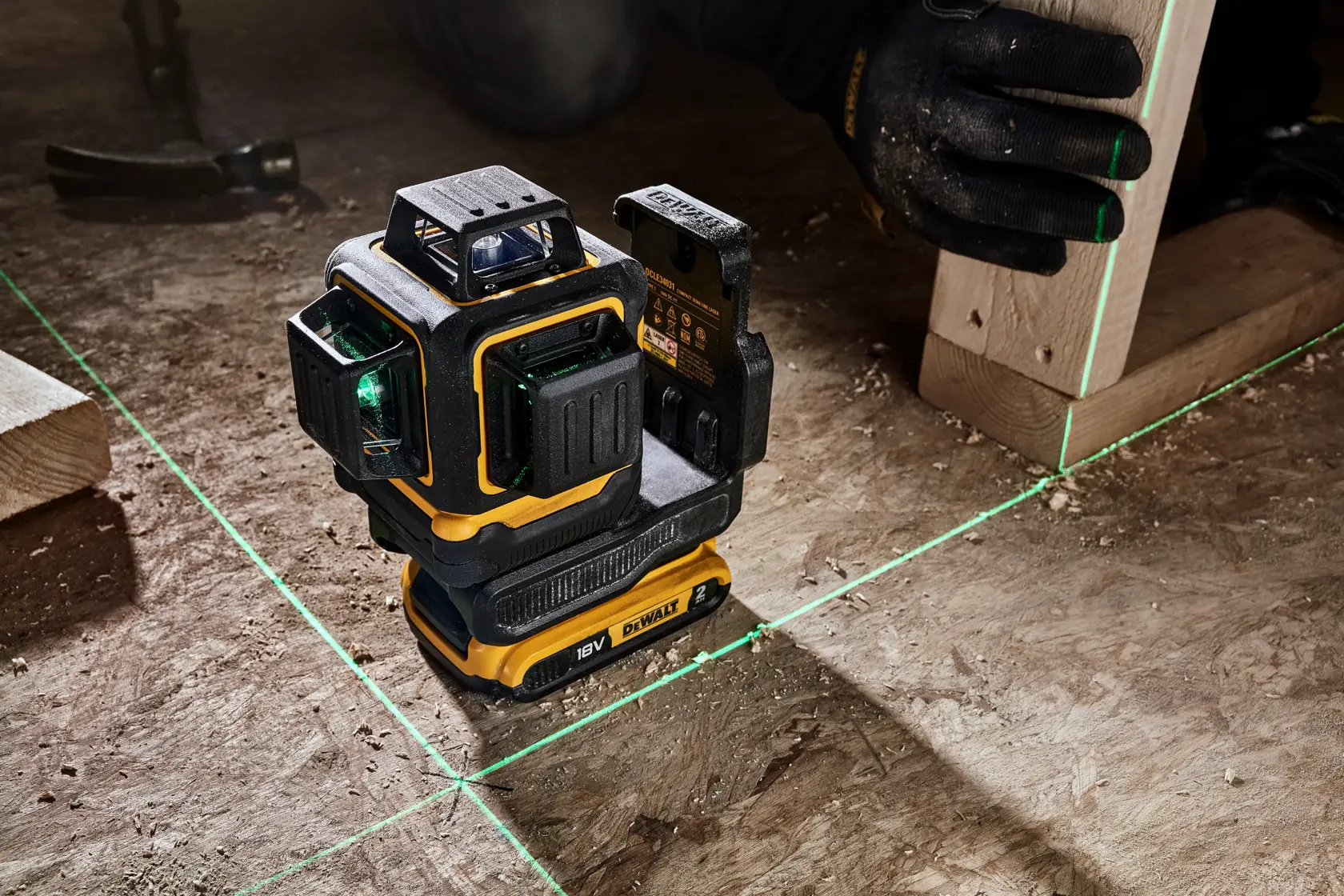 Una herramienta de nivel láser DEWALT proyecta líneas láser verdes sobre un suelo de madera, utilizada para la alineación precisa durante la construcción. Una persona con guantes sostiene una viga de madera junto a las líneas láser.