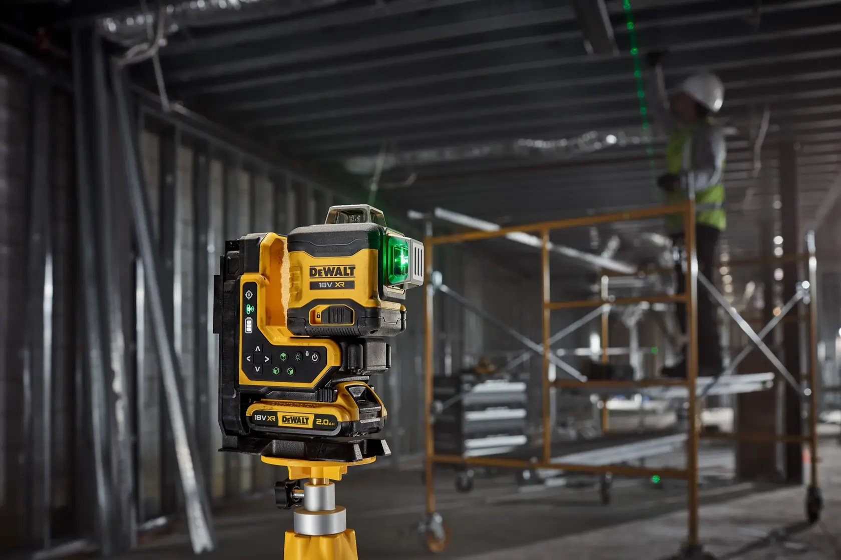 DEWALT Laser mit grünem Strahl auf Stativ an Baustelle, Arbeiter und Gerüst im Hintergrund.