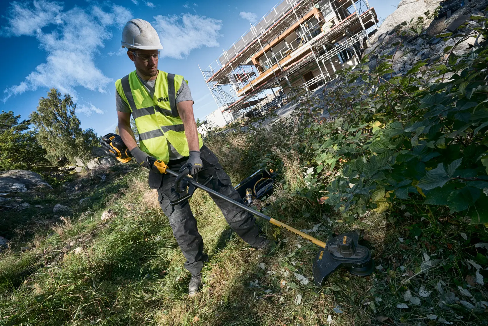 Trabajador usa cortabordes DEWALT al aire libre, con protección cerca de obras y andamiaje.