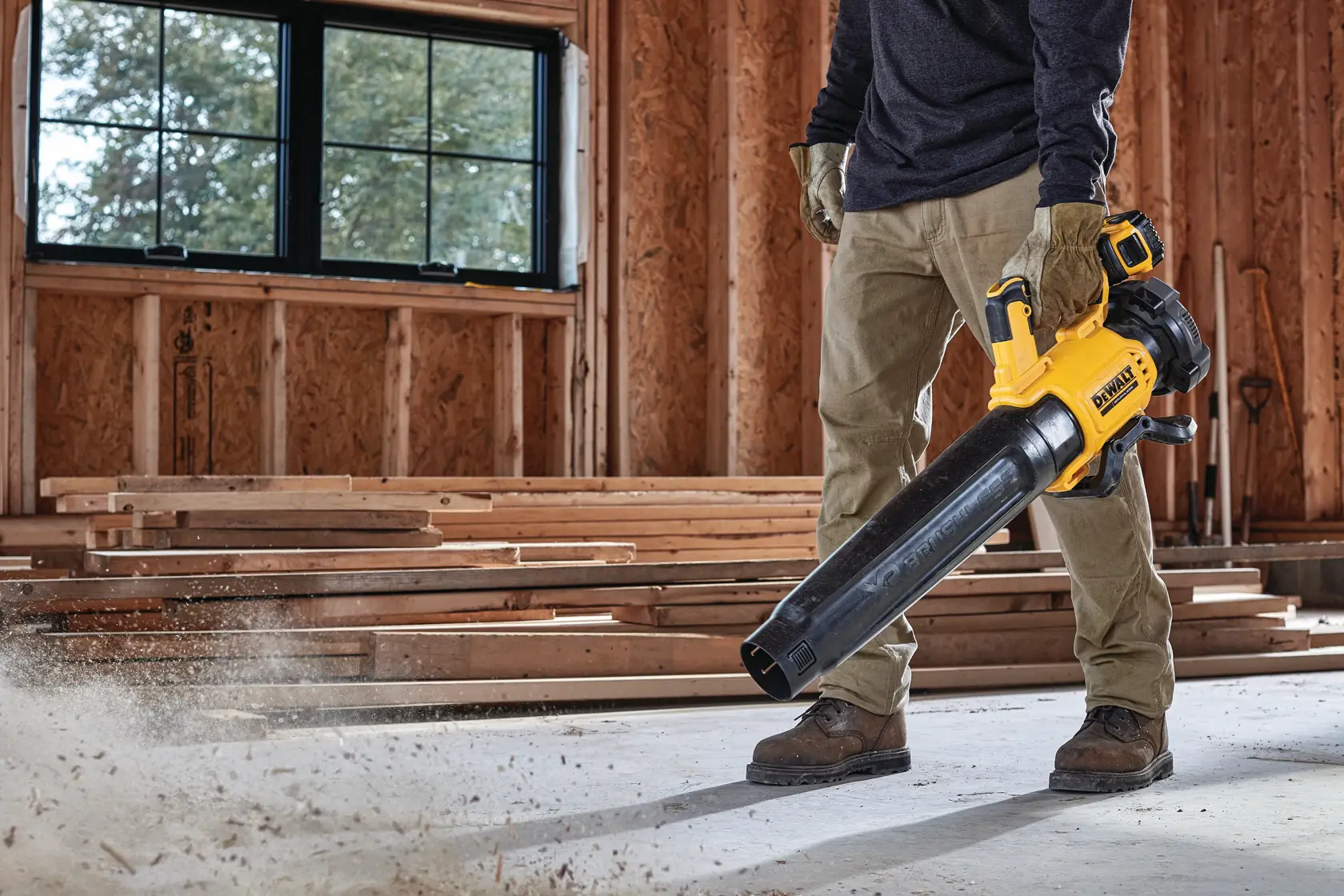 Persona usando soplador DEWALT inalámbrico para limpiar polvo en sala con estructura de madera en obra.