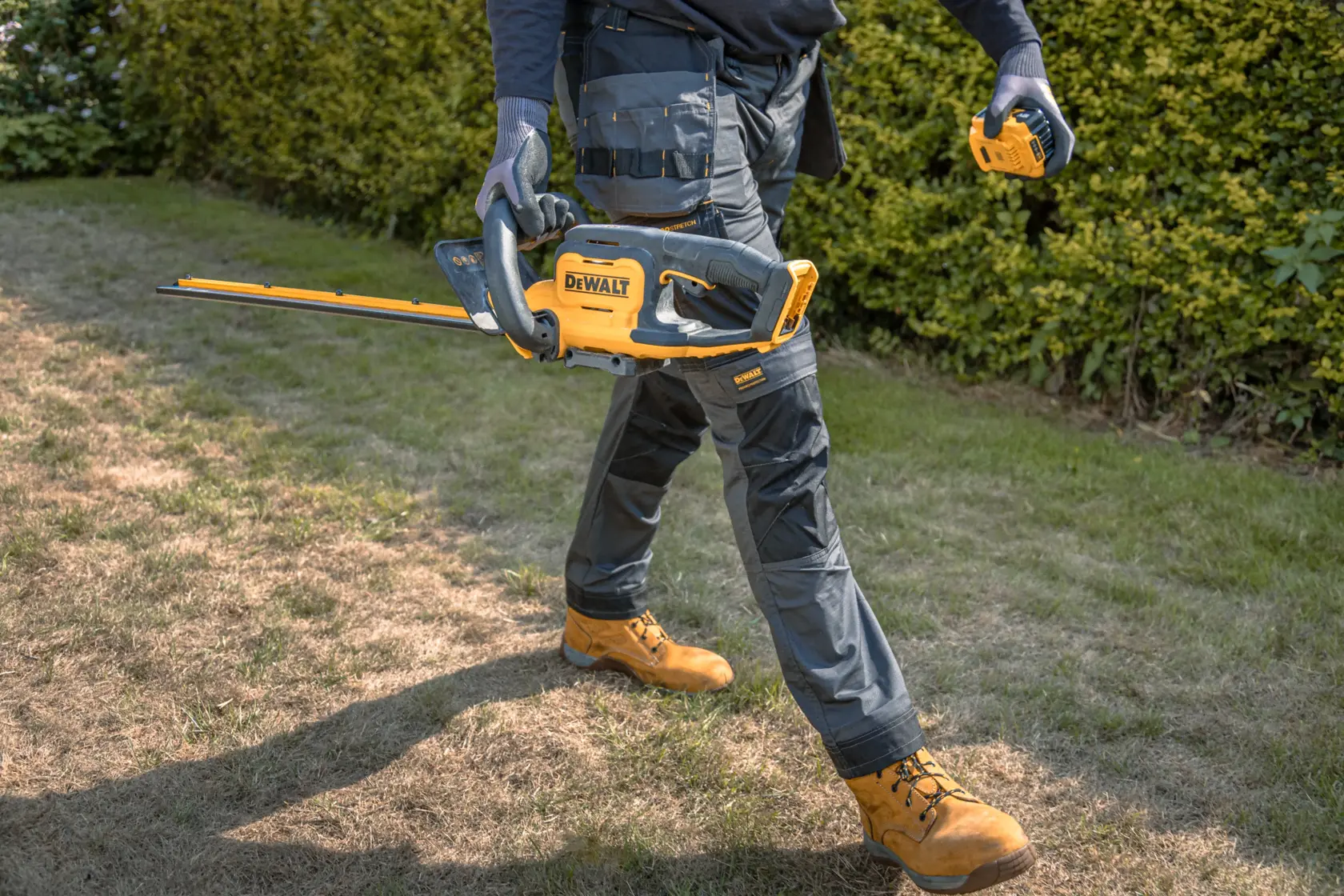 Una persona con guanti da lavoro e abbigliamento protettivo tiene in mano un tagliasiepi a batteria DEWALT giallo e nero e cammina su un prato accanto a dei cespugli.