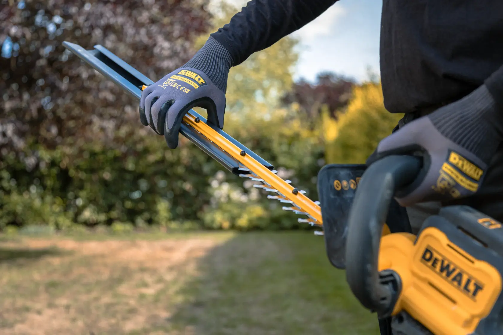 Persona usa un tagliasiepi senza fili DEWALT in giardino indossando guanti protettivi.