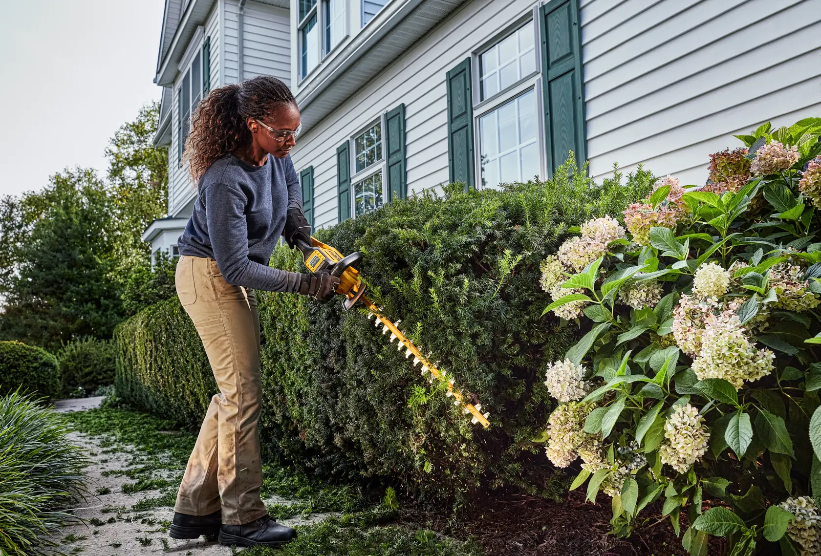 Persona usando una recortadora de setos DEWALT para recortar arbustos frente a una casa con revestimiento blanco y persianas verdes.