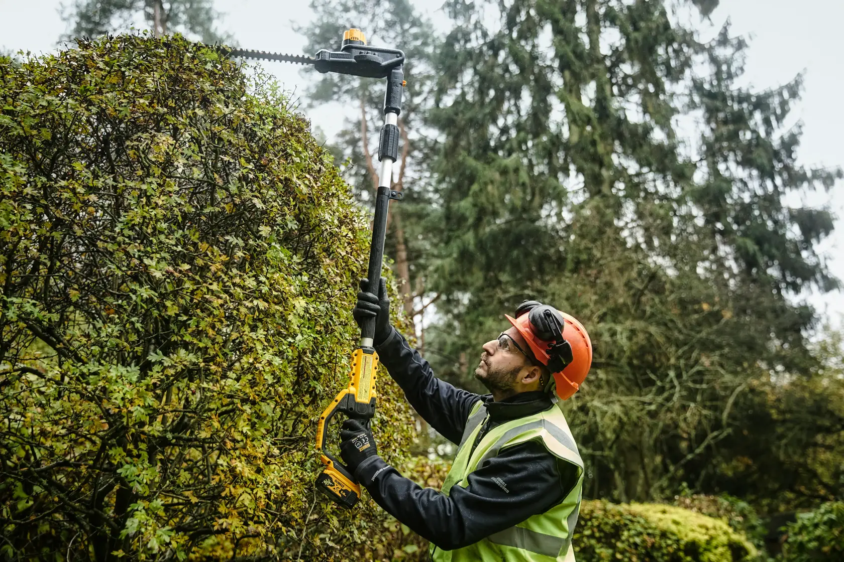Trabajador recorta seto alto con DEWALT DCMPH566N inalámbrico, usando equipo de seguridad.