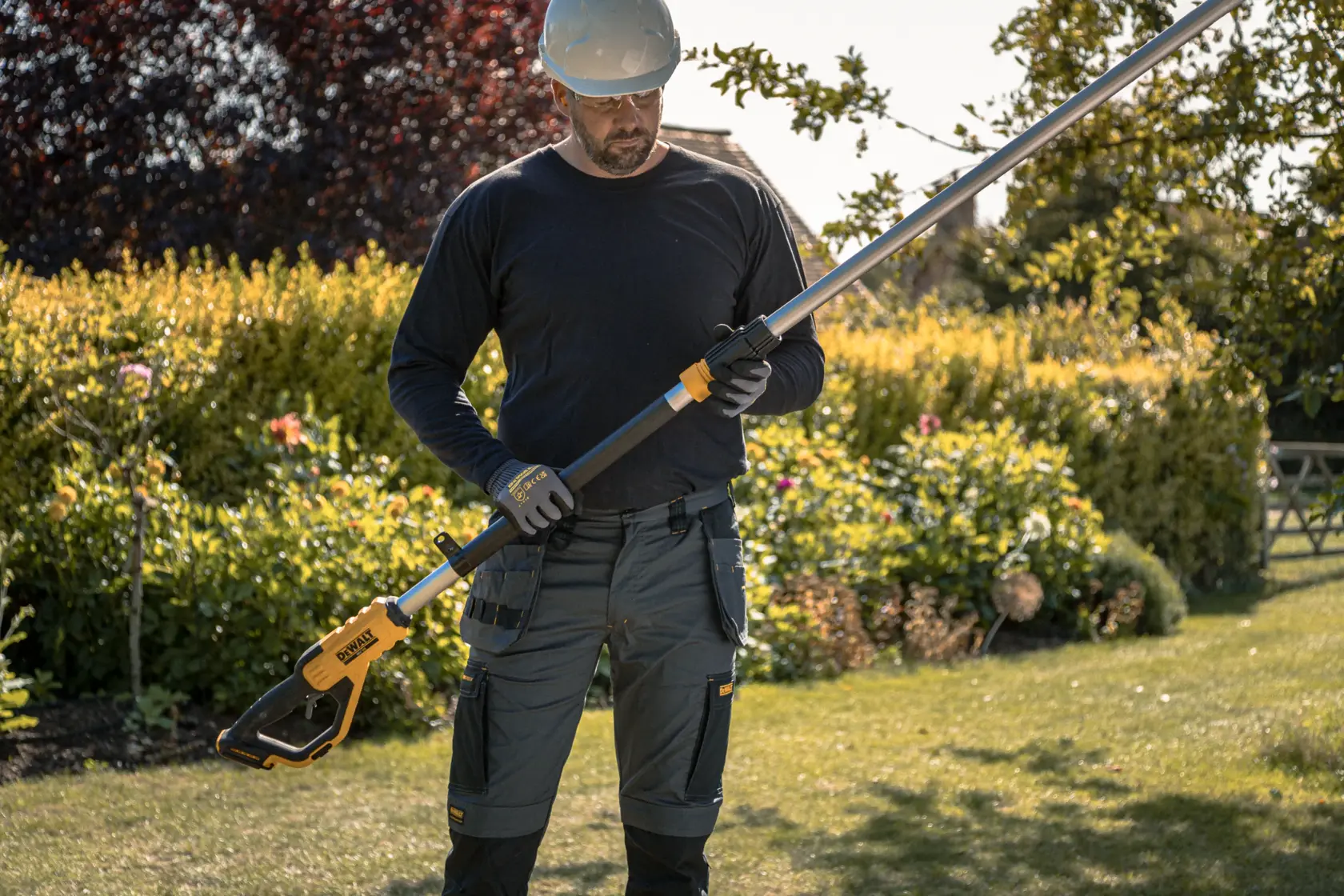 Persona con una sega a palo DEWALT senza fili all'aperto, piante di giardino sullo sfondo.