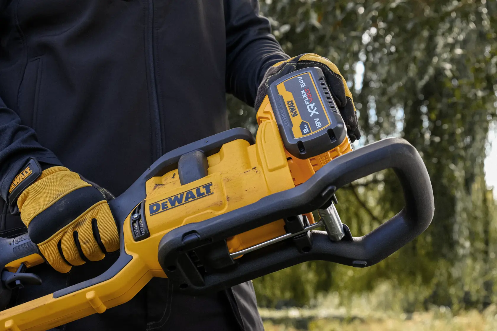 Behandschuhte Hände halten DEWALT Akku-Kettensäge mit sichtbarer XR FLEXVOLT 18V Batterie im Freien.