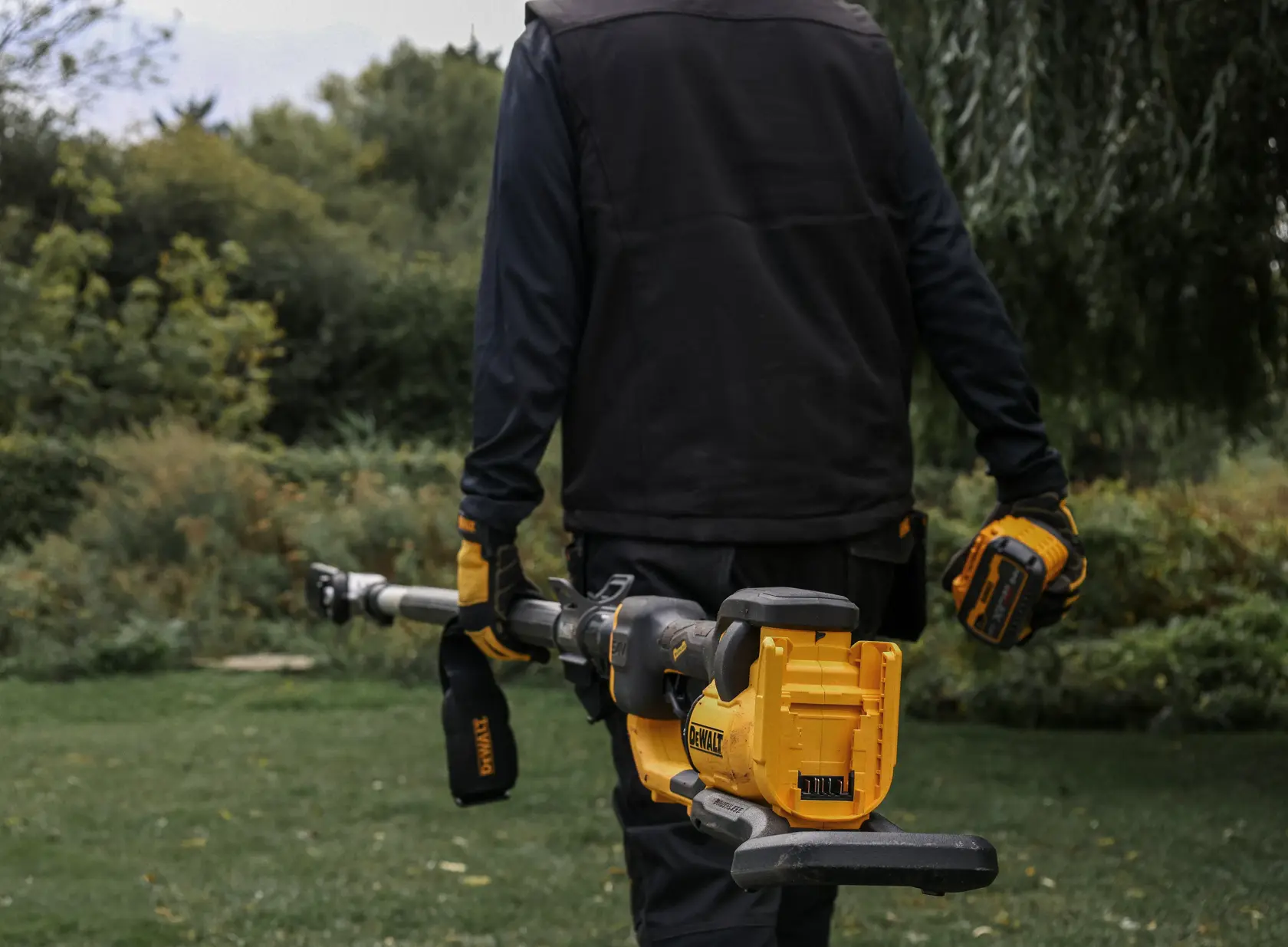 Person hält gelben DEWALT Akku-Astschneider im Garten, trägt Handschuhe und Arbeitskleidung.
