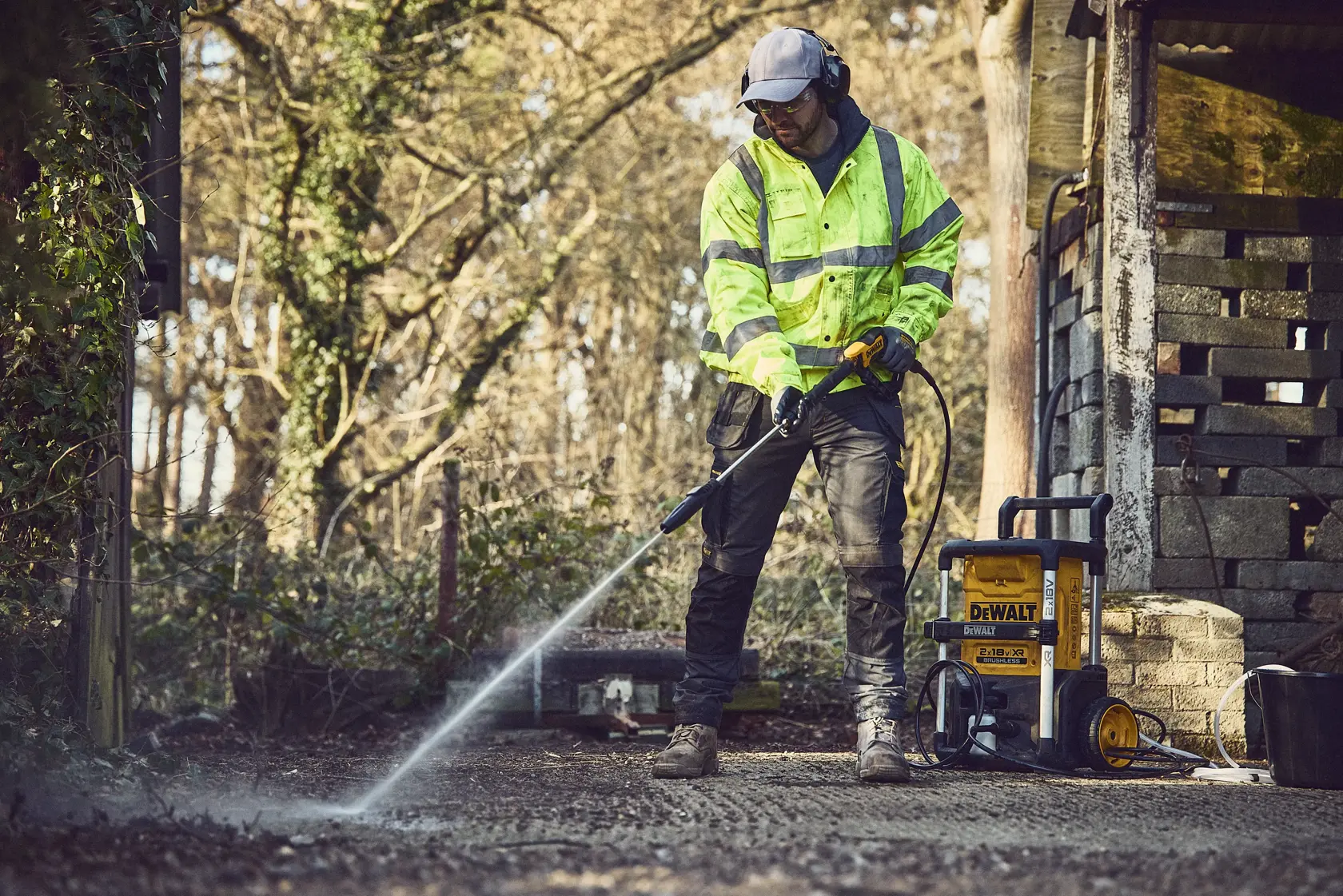 Persona usa una hidrolavadora DEWALT en exterior sobre pavimento junto a un cobertizo, equipada con protección.