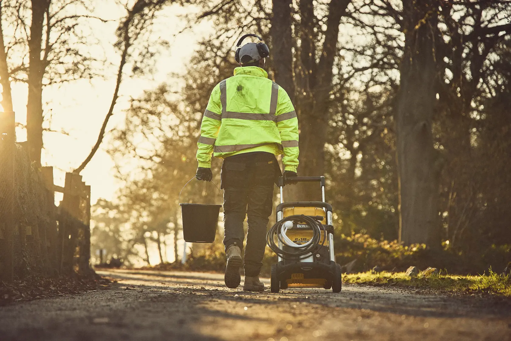 Persona con ropa de seguridad lleva una hidrolimpiadora DEWALT y un balde por un sendero rural al atardecer.
