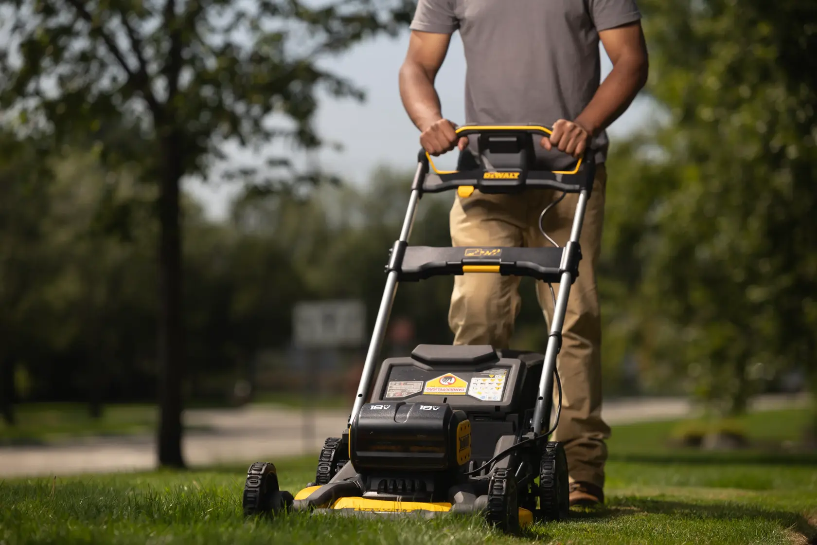 A person is pushing a DEWALT cordless lawn mower outdoors on a grassy lawn. The focus is on the lawn mower, showing its robust design and battery compartment.