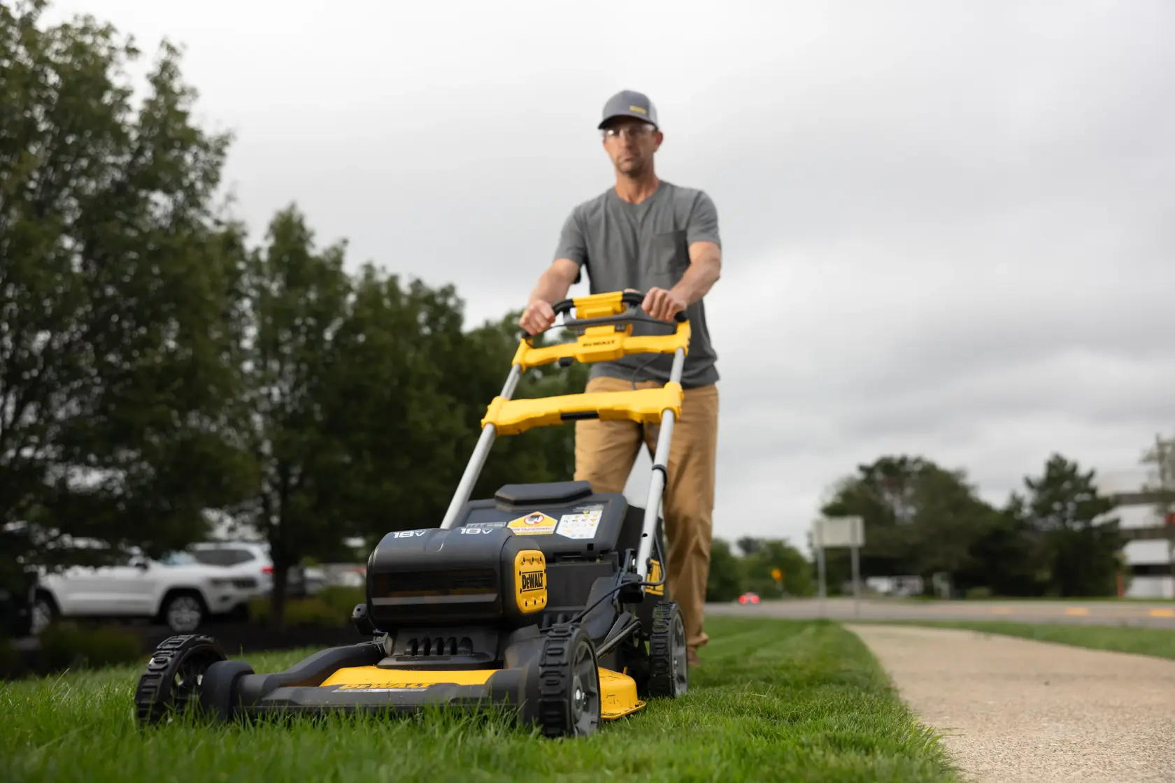Una persona empuja una cortadora de césped DEWALT sobre el césped junto a una acera, con árboles y autos estacionados al fondo. El rostro de la persona está difuminado.
