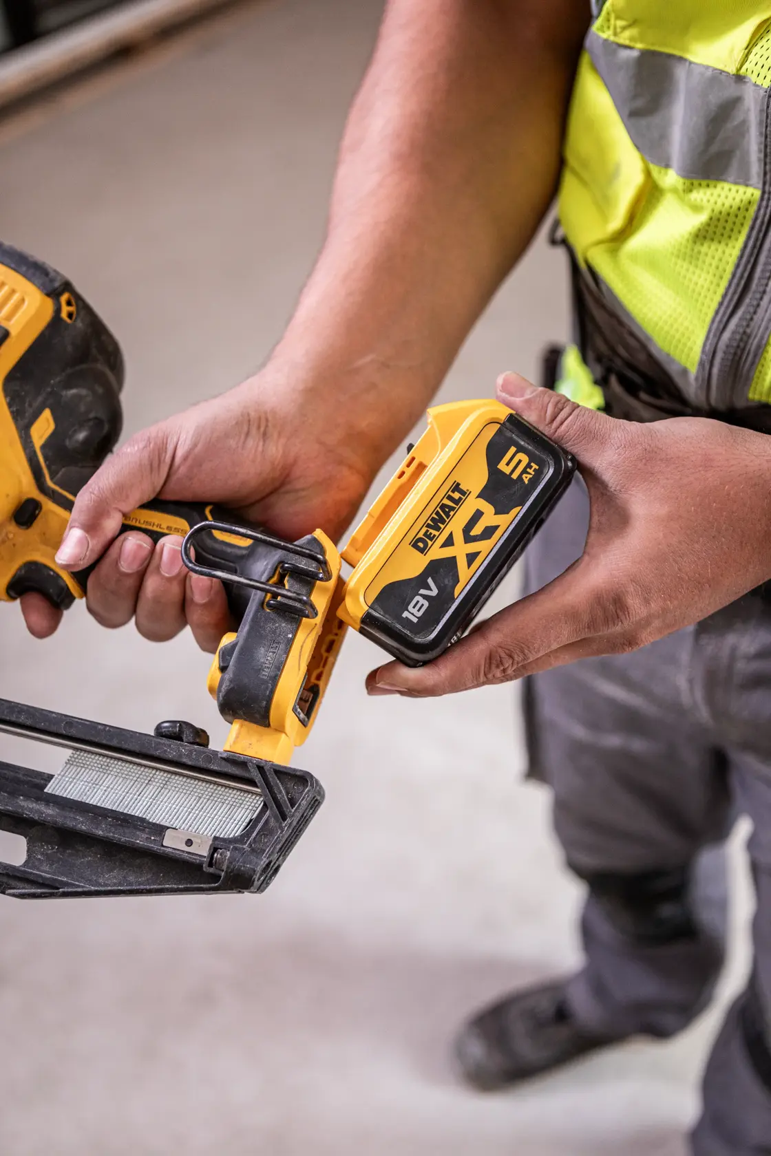 Person holding a DEWALT cordless tool with an 18V XR 5Ah battery, wearing work clothes indoors.