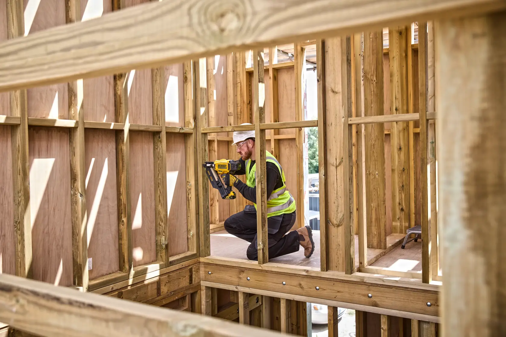 Trabajador utiliza herramienta DEWALT para fijar marcos de madera en una obra, con equipo de seguridad y de rodillas.