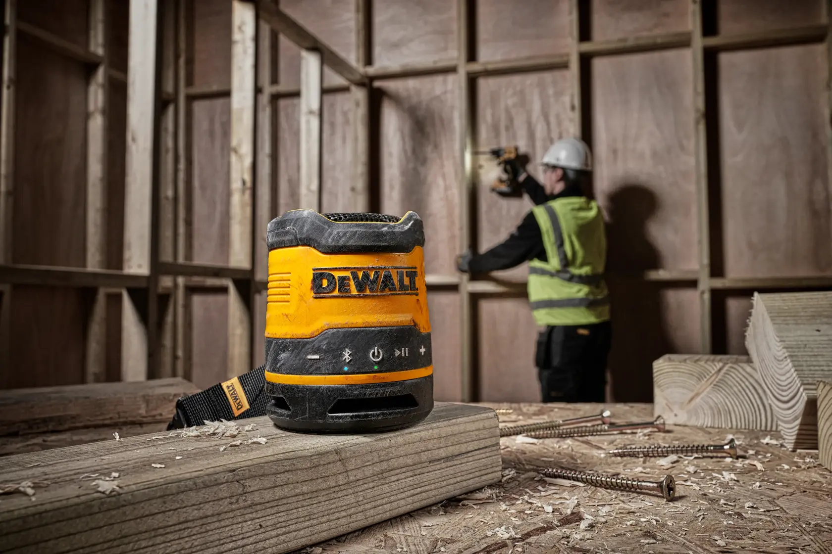 A DEWALT DCR009 portable speaker placed on a wooden beam at a construction site, with a worker in safety gear operating a tool in the background.
