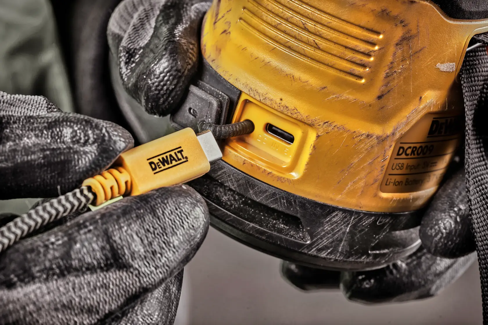 Close-up of a person wearing black gloves plugging a yellow DEWALT USB-C charging cable into a DEWALT DCR009 device. The device has visible scratches and a product label.