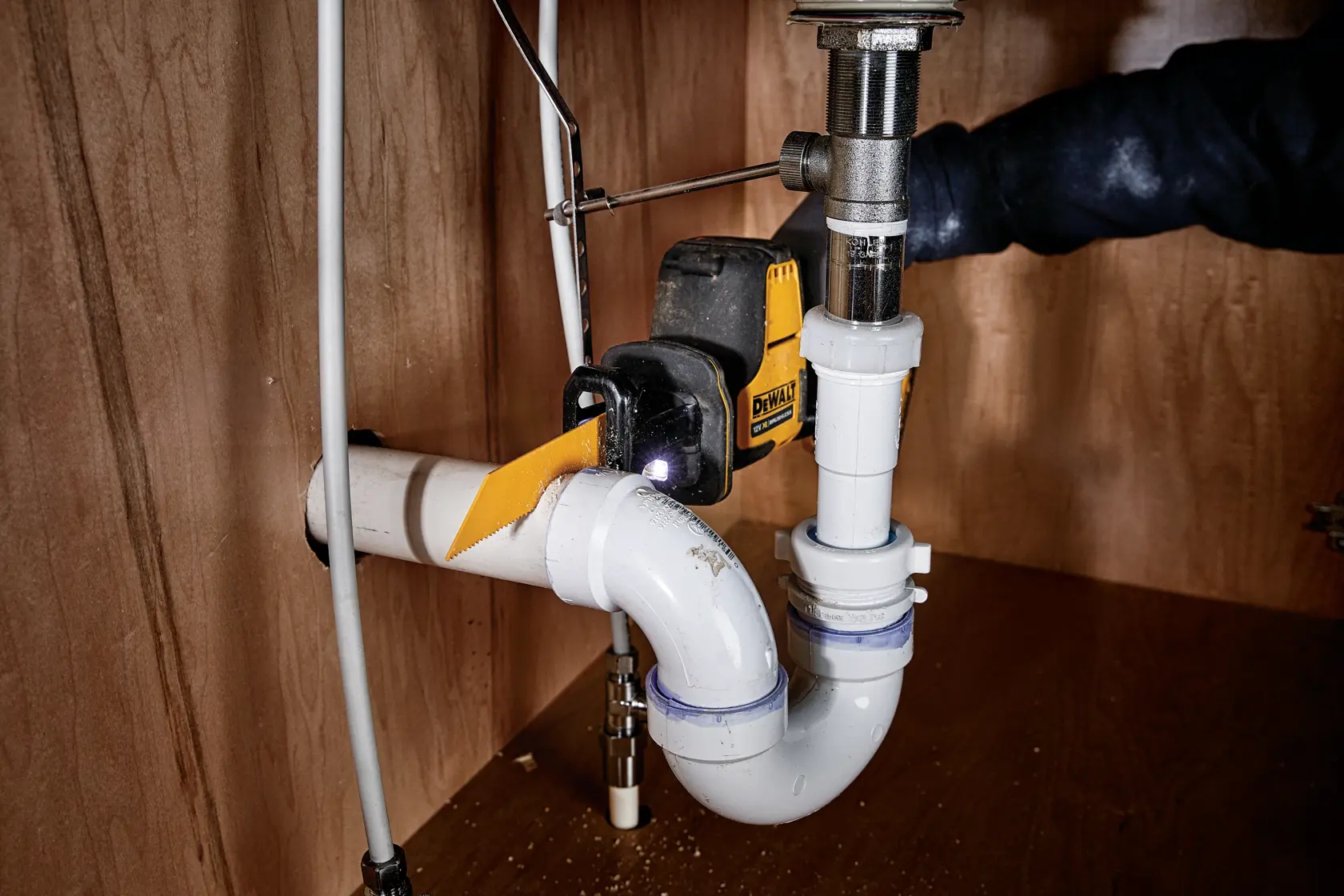 Herramienta DEWALT cortando tubería de PVC blanca bajo un fregadero en mueble de madera.