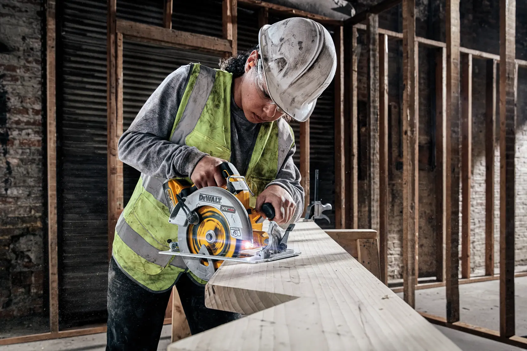 Eine Person mit Sicherheitsweste und Schutzhelm verwendet eine DEWALT DCS573H2T Akku-Kreissäge, um ein großes Holzbrett auf einer Baustelle zu schneiden.