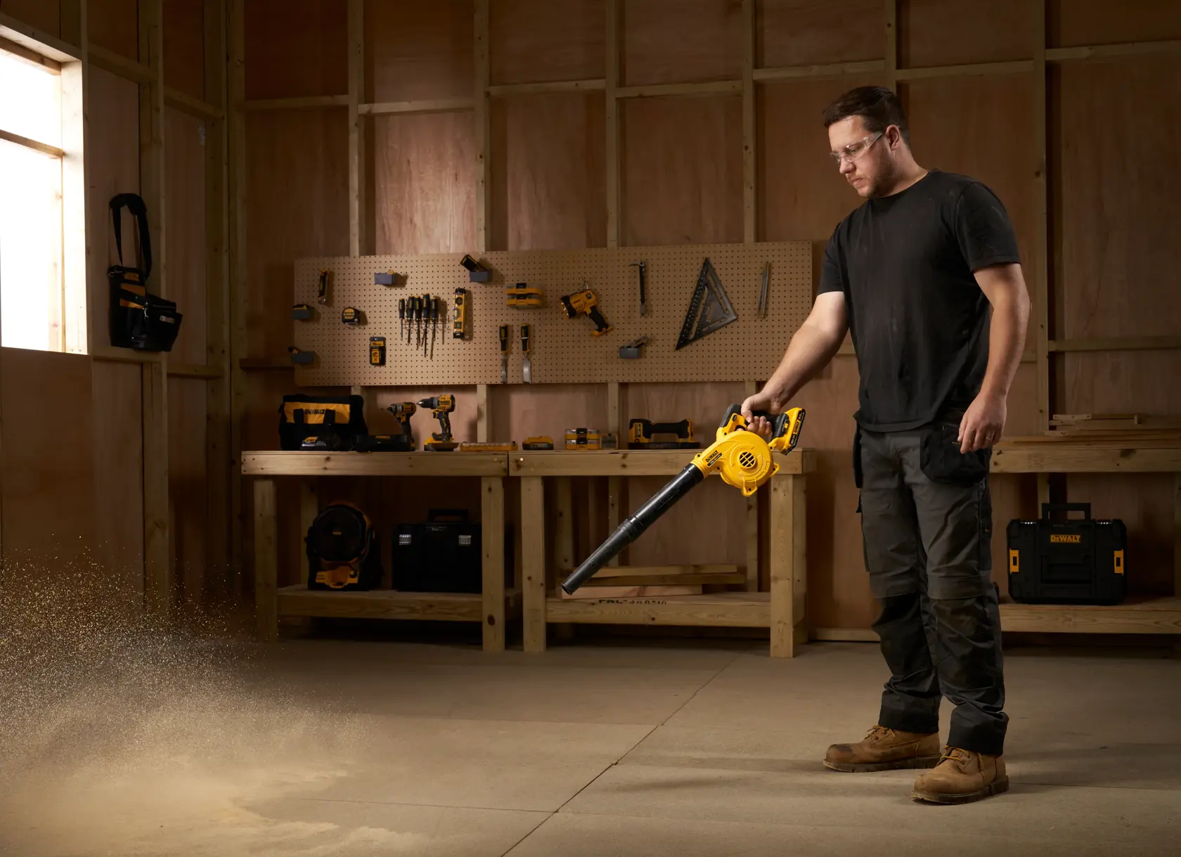 Persona usa soplador DEWALT DCV100 para limpiar polvo en taller con herramientas y banco de madera.